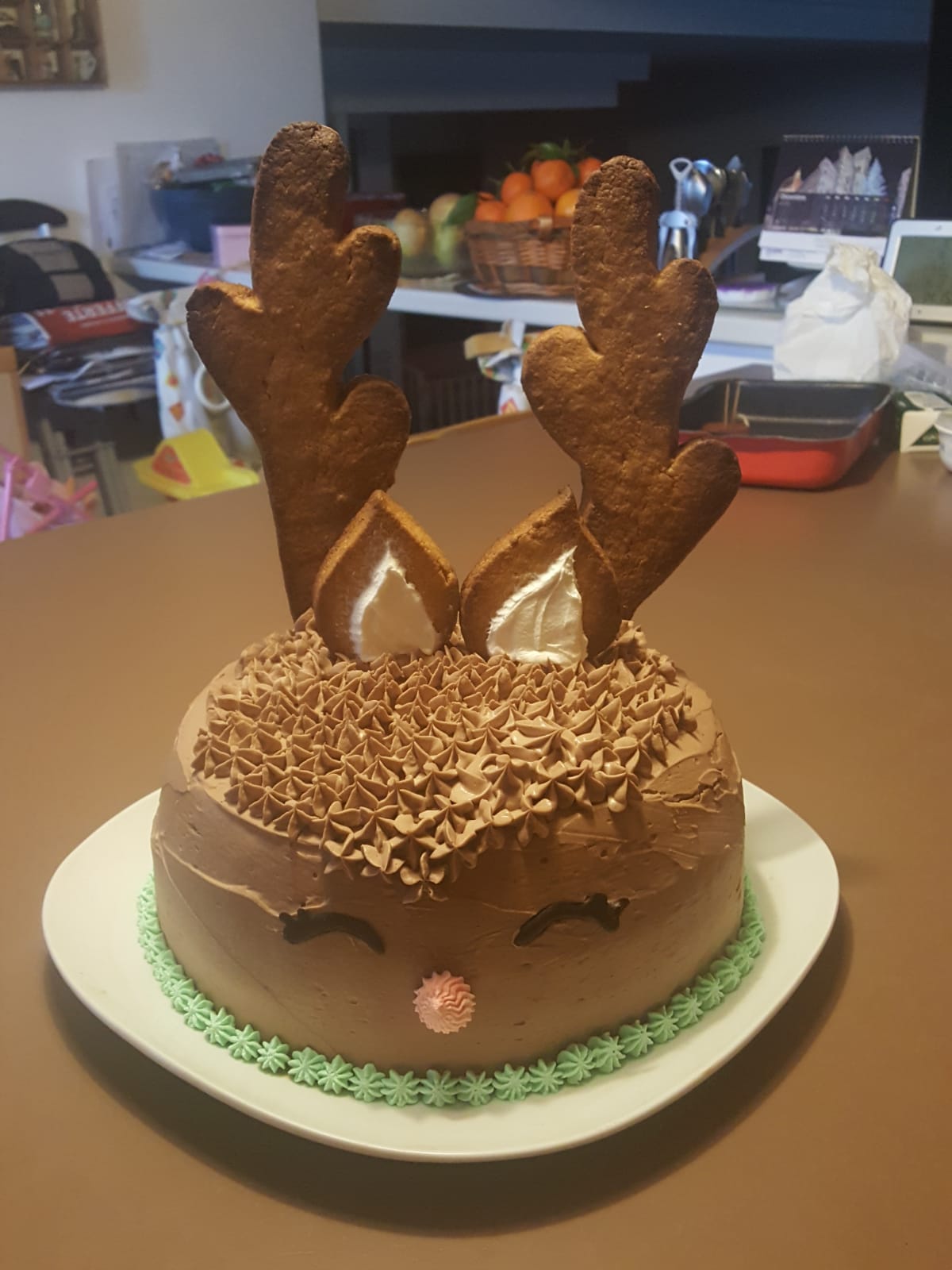 A round cake decorated as a reindeer with chocolate icing, cookie antlers, piped ears, closed eyes, and a pink nose, sitting on a white plate on a kitchen counter.