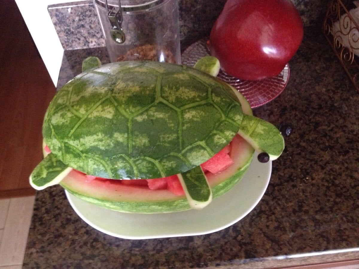 A watermelon carved to look like a turtle, with the top resembling a turtle shell, and the inside filled with watermelon pieces. The turtle’s head and legs are made from watermelon and fruits.