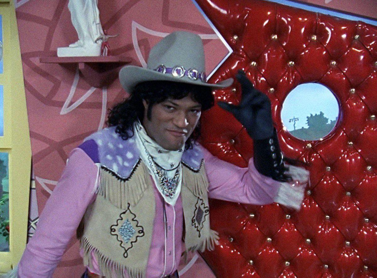 A person in a cowboy outfit with a light hat, pink shirt, and beige vest poses in a colorful, retro room with a red cushioned wall and a circular window.
