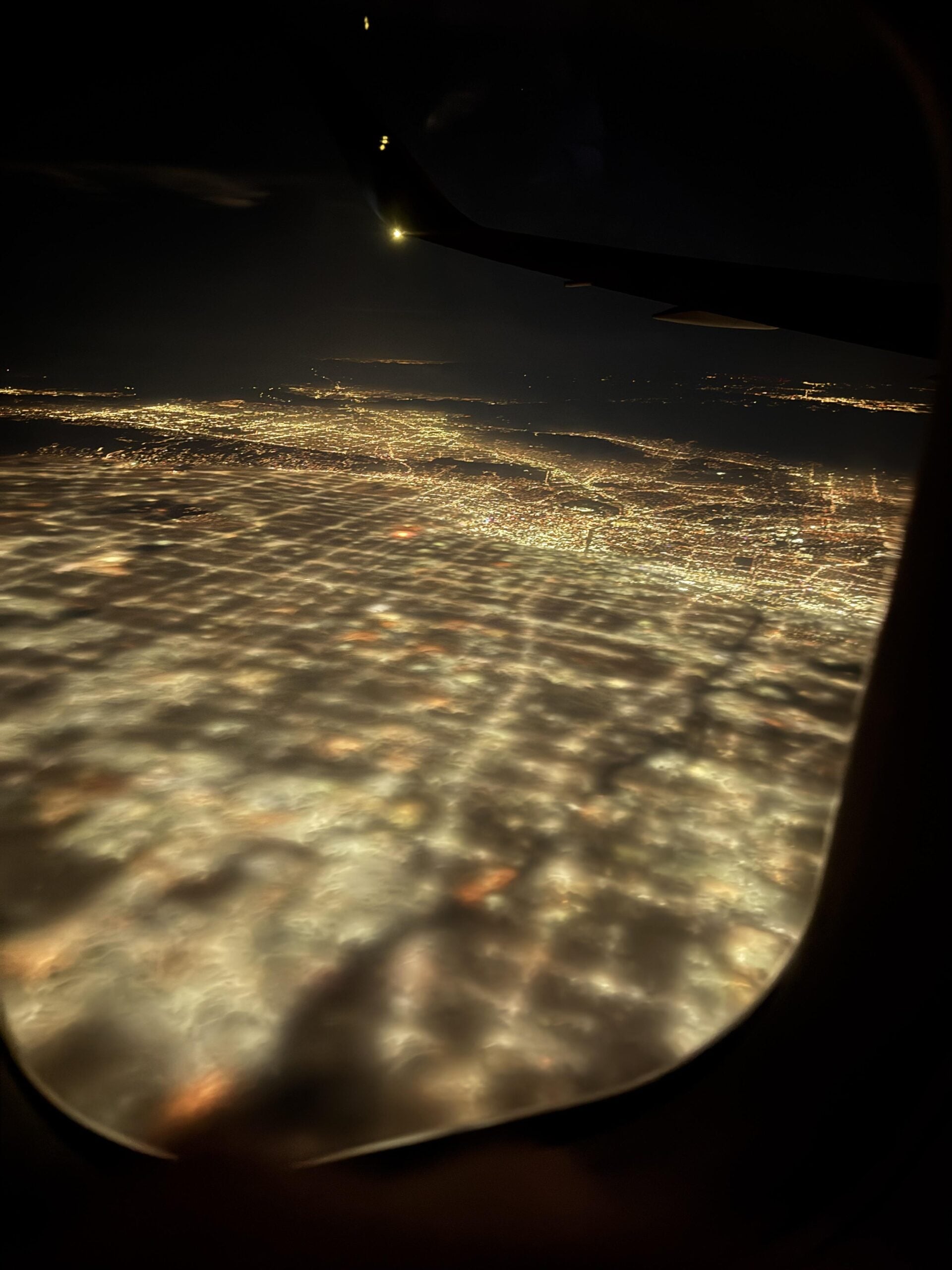 Aerial view from an airplane window at night, showing a glowing grid of city lights below partially covered by clouds, with part of the plane’s wing visible.