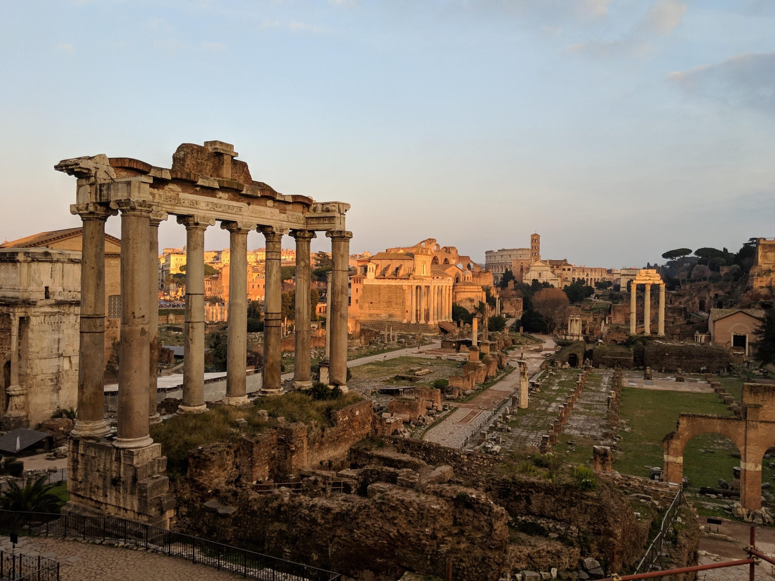 Ancient Roman ruins bathed in warm sunlight, featuring tall stone columns and remnants of buildings with grassy and stone pathways, set against a partly cloudy sky.