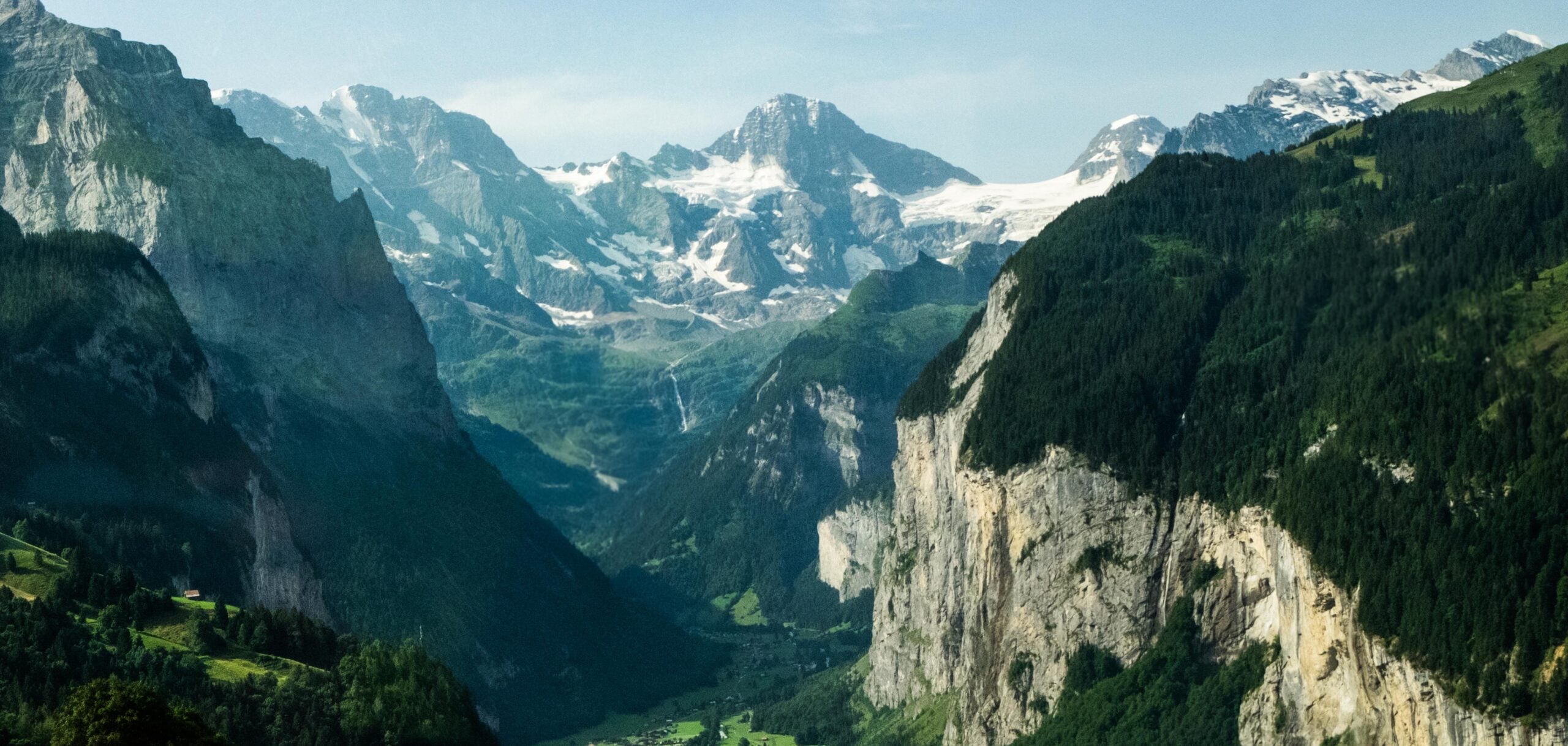 A lush green valley surrounded by steep cliffs and forested mountains, with snow-capped peaks in the background under a clear blue sky.