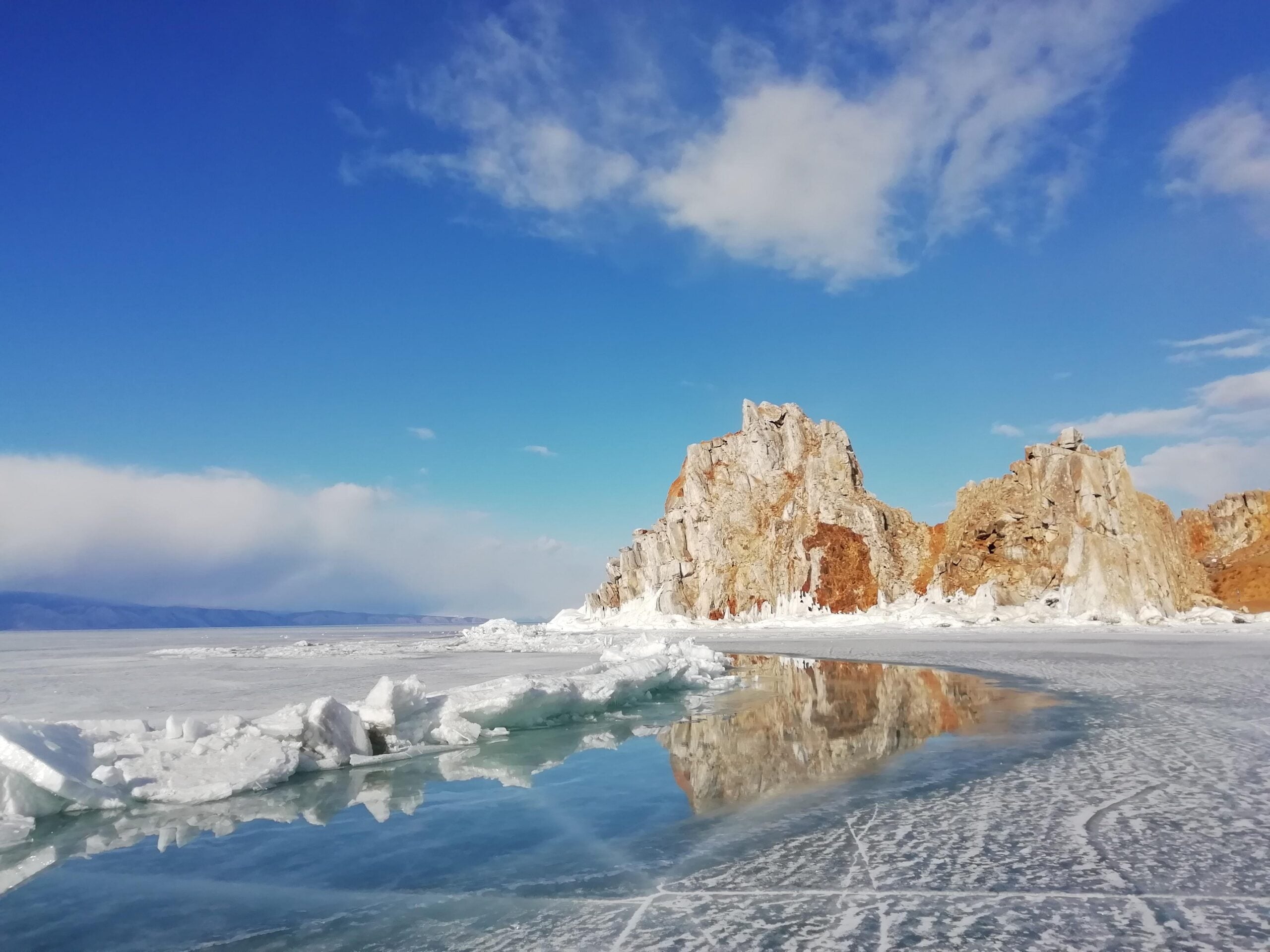 Rocky cliffs rise above a frozen lake with patches of clear blue ice reflecting the sky, under scattered clouds on a bright day.