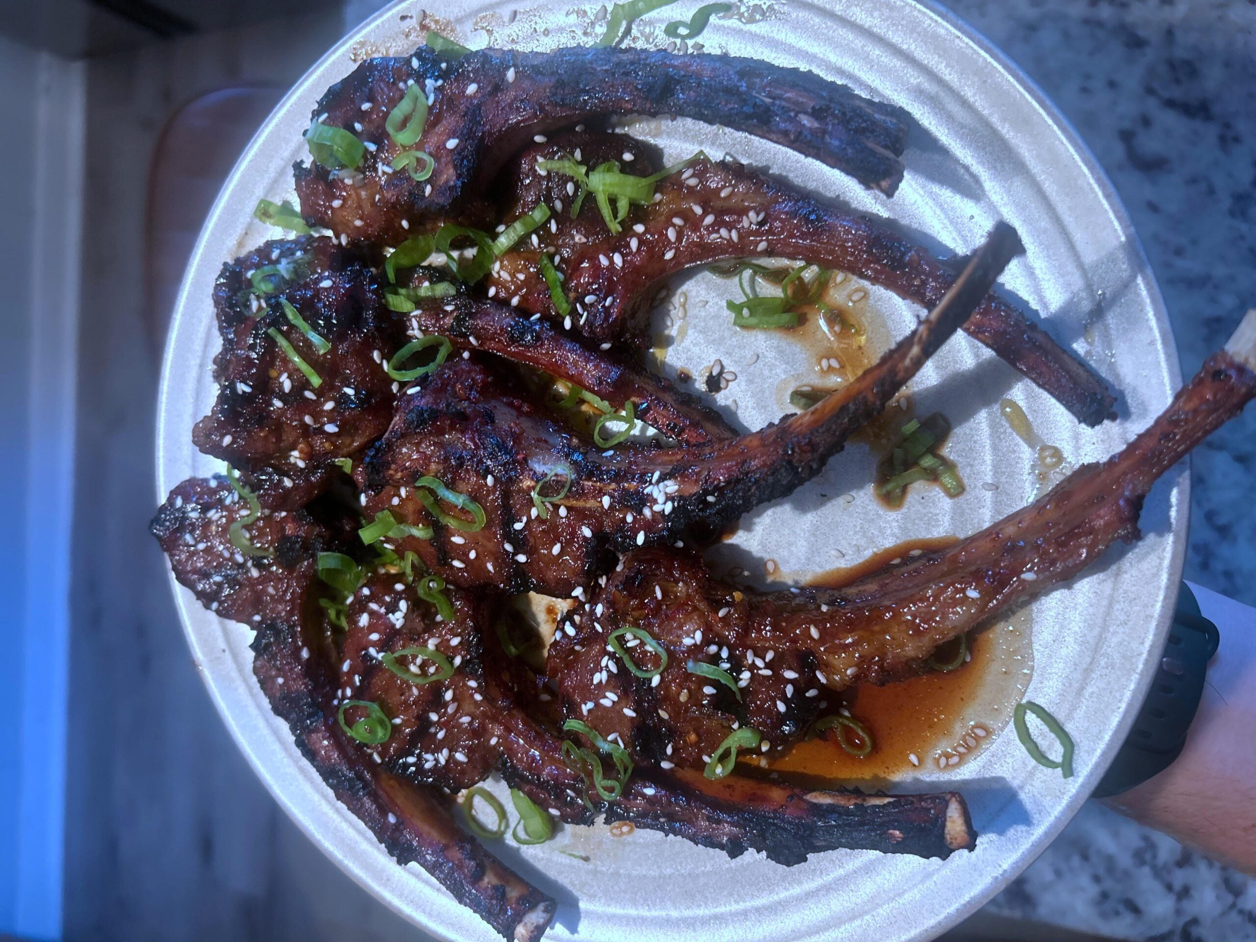 A plate of grilled lamb chops glazed with sauce, garnished with sliced green onions and sesame seeds, sits on a countertop.