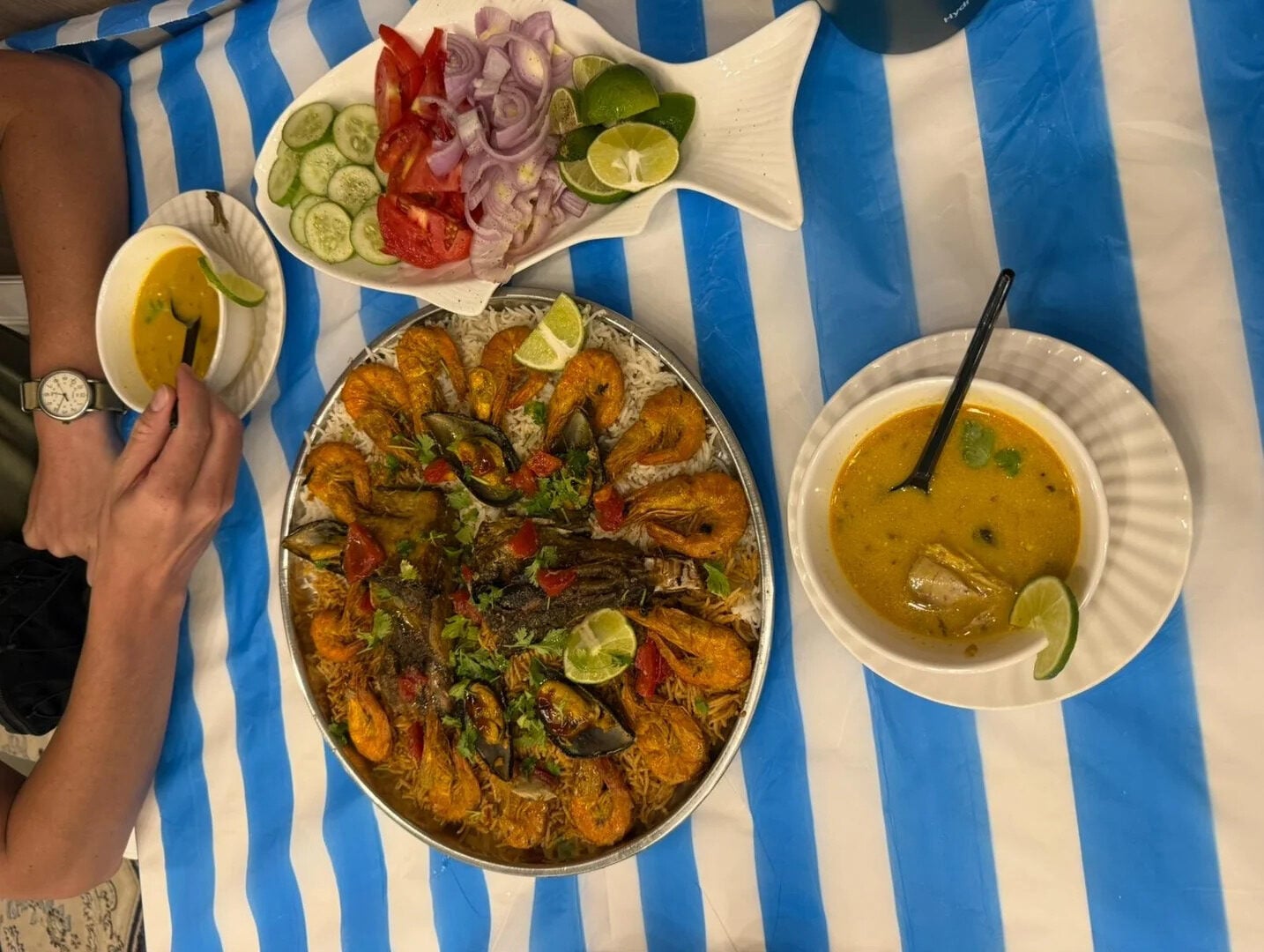 A table filled with Amazing Food People Ate While On Vacation: prawn biryani, a bowl of yellow curry, and a fish-shaped plate of sliced cucumbers, tomatoes, and onions. A hand holds a small bowl of curry over the vibrant spread. The tablecloth is blue and white striped.