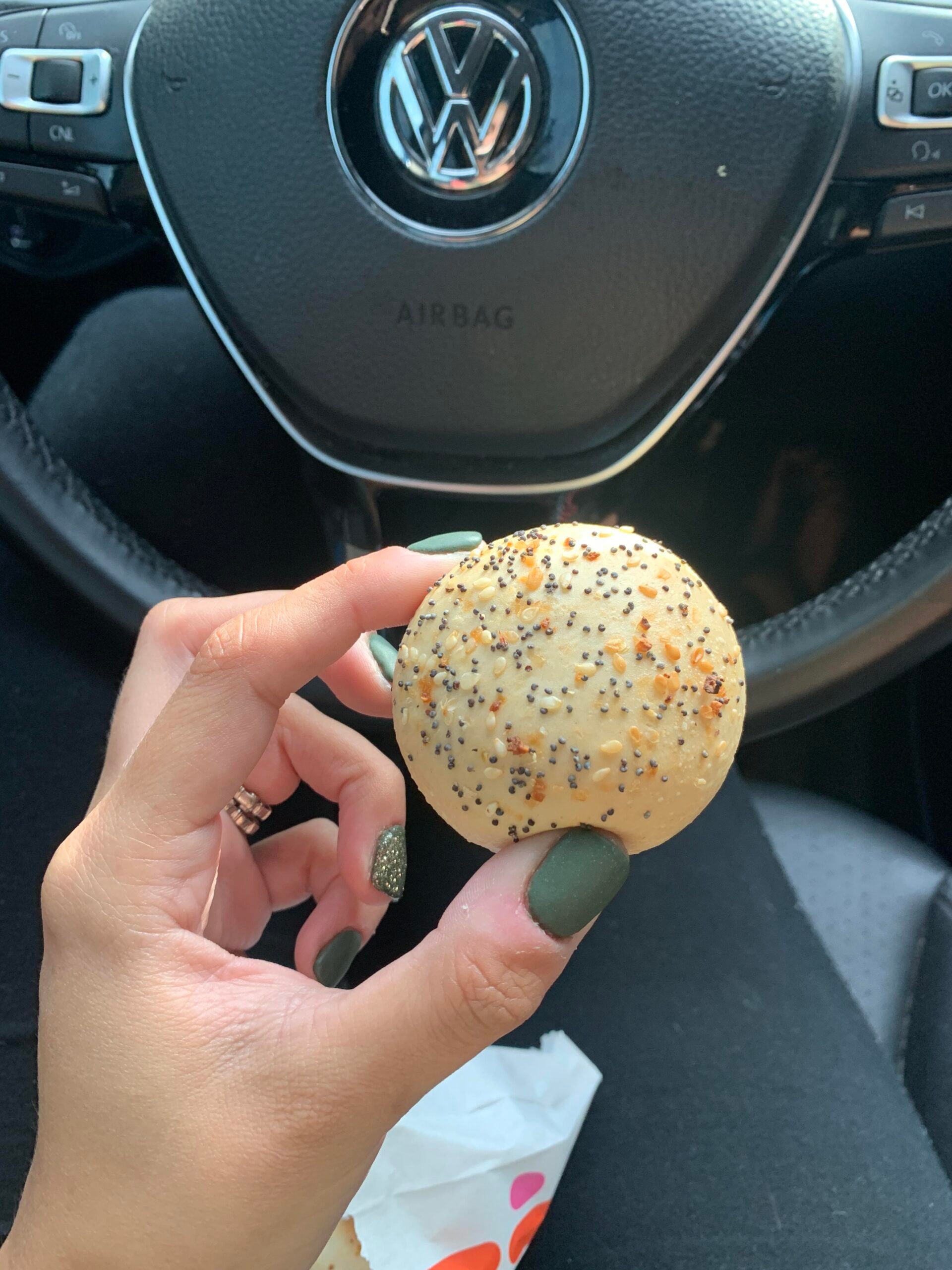 A hand with green-painted nails holds a round, seeded bagel bite inside a car, with a Volkswagen steering wheel in the background.