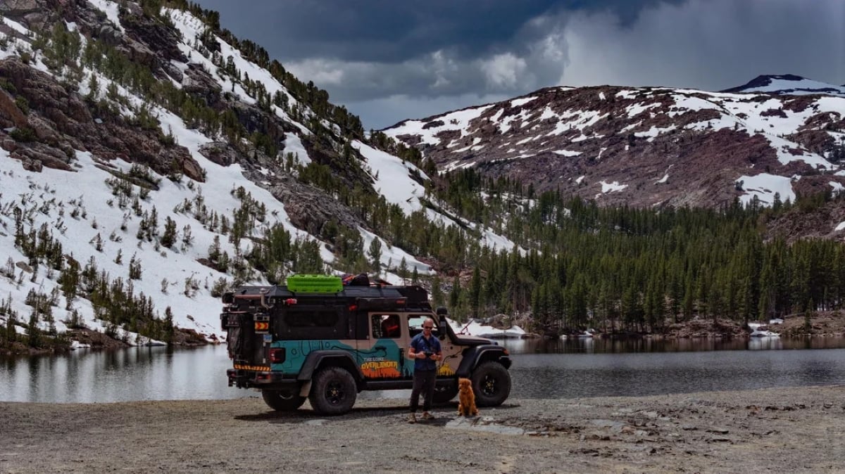 A person with a dog stands beside an off-road vehicle near a lake, surrounded by snow-capped mountains and pine trees under a cloudy sky.