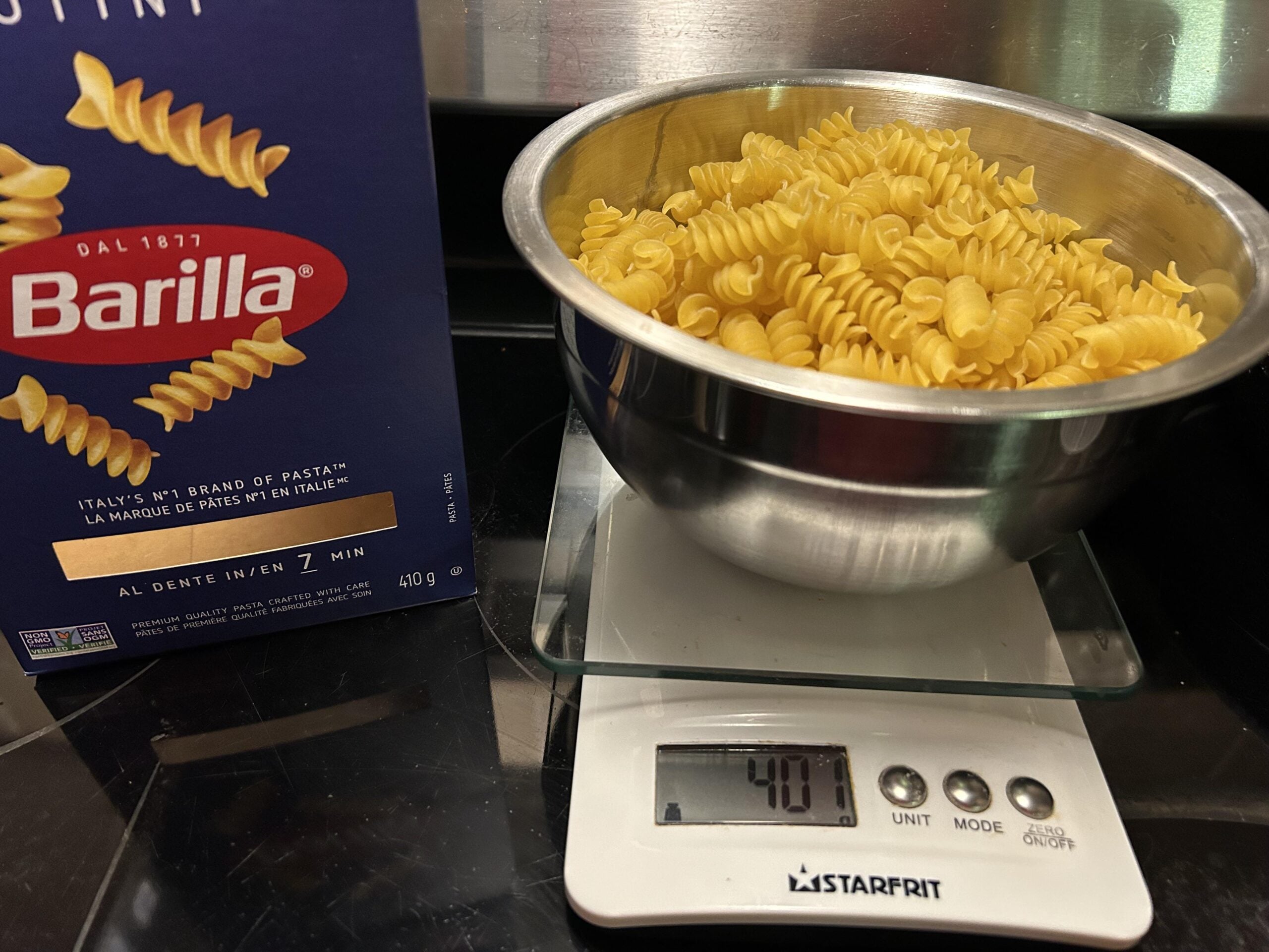 A metal bowl filled with uncooked rotini pasta sits on a digital scale showing 401 grams, next to a Barilla pasta box on a black kitchen counter.