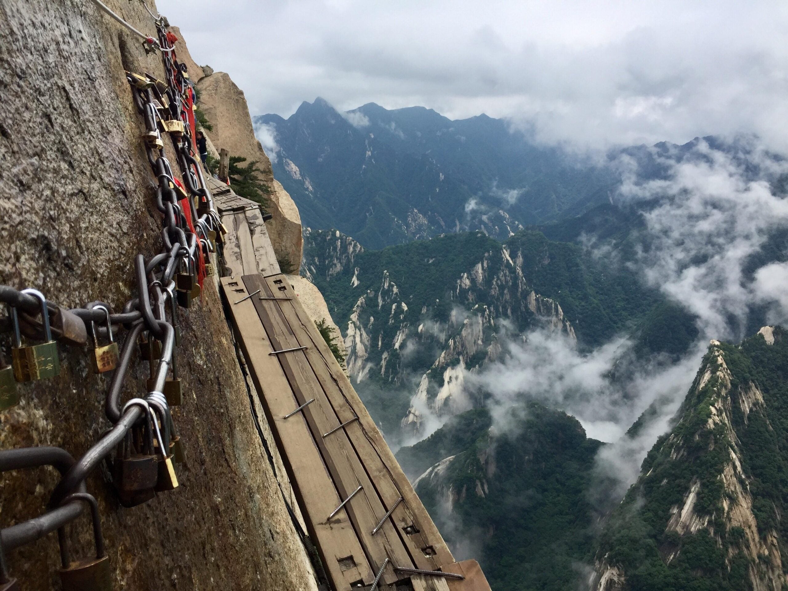 A narrow wooden walkway clings to a steep mountain cliff, secured by chains with padlocks. Mist and clouds drift among dramatic, rugged peaks in the background.
