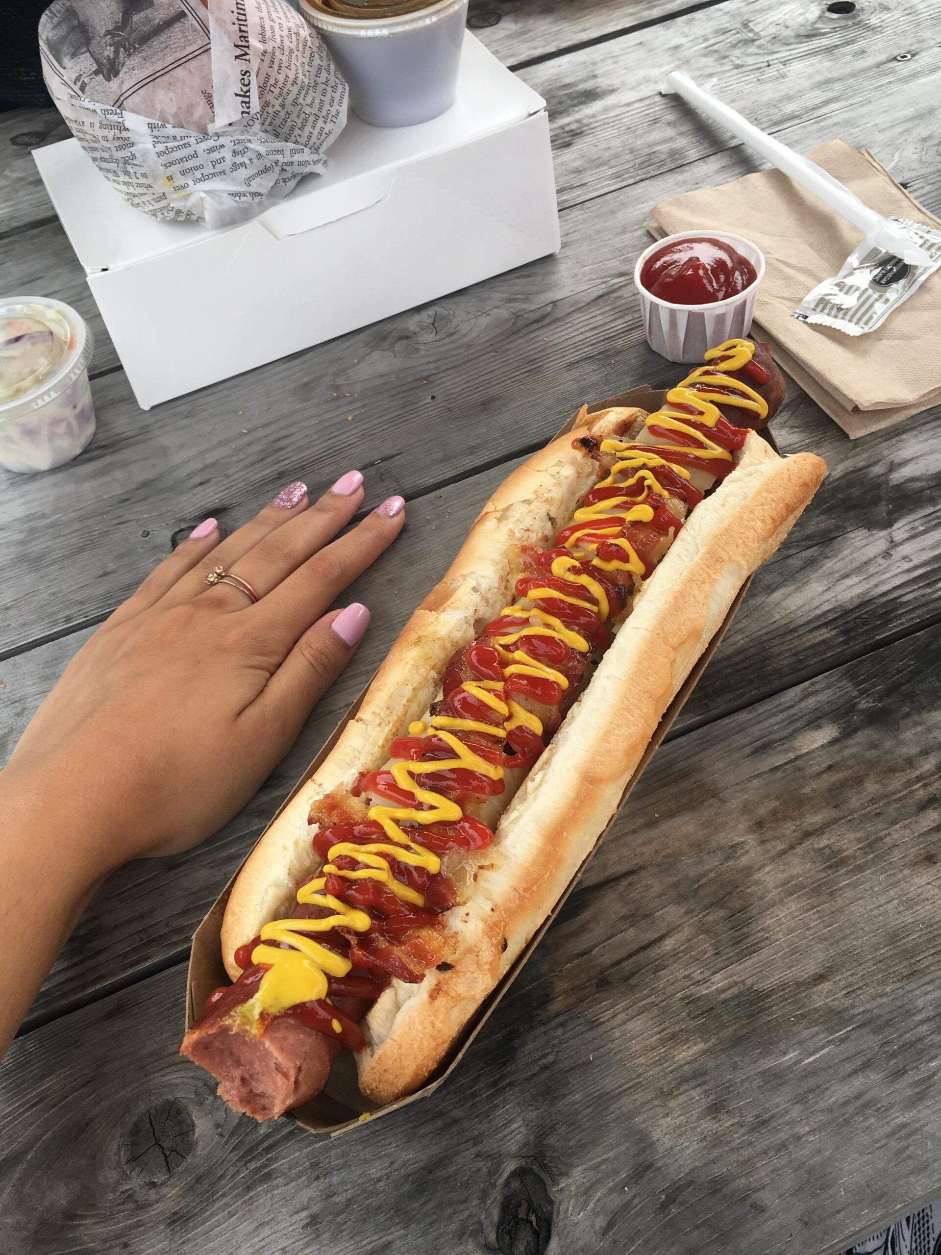 A hand next to a very large hot dog topped with ketchup and mustard on a wooden table, with a fork, napkin, ketchup cup, and other food items nearby.