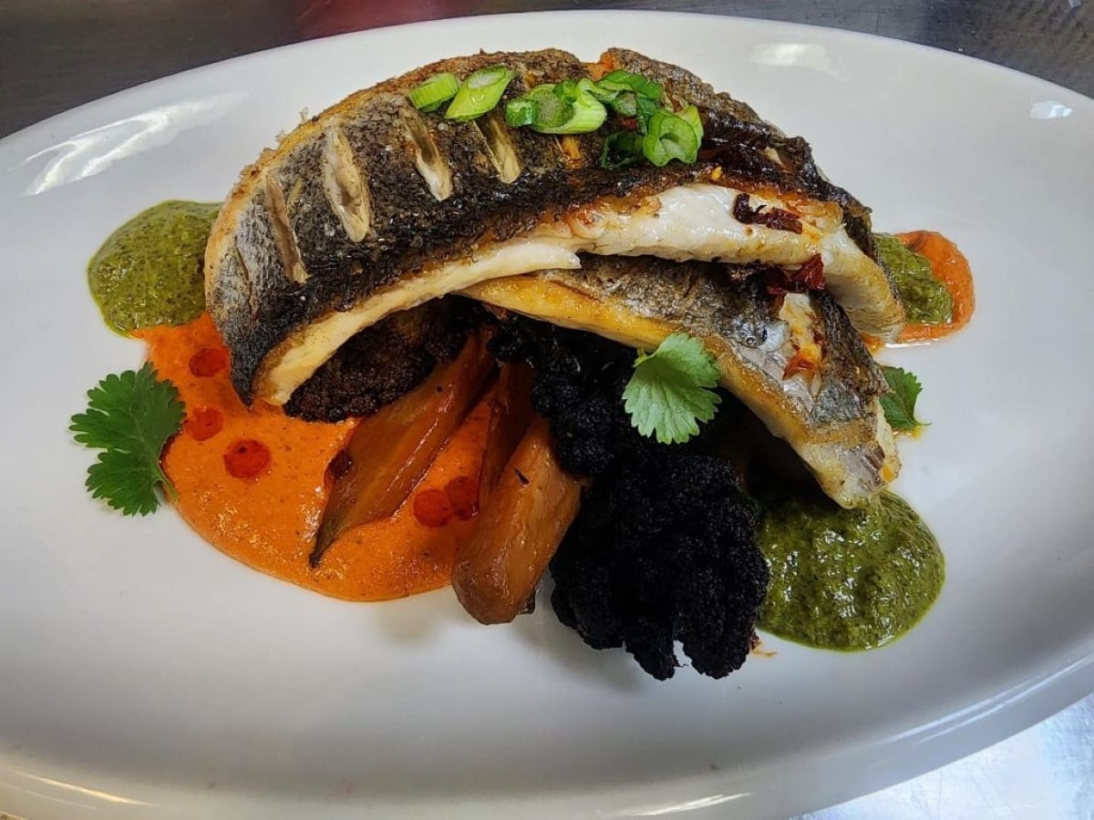 Grilled fish fillet served on roasted carrots and vegetables, garnished with chopped green onions and cilantro, plated with red and green sauces on a white dish.