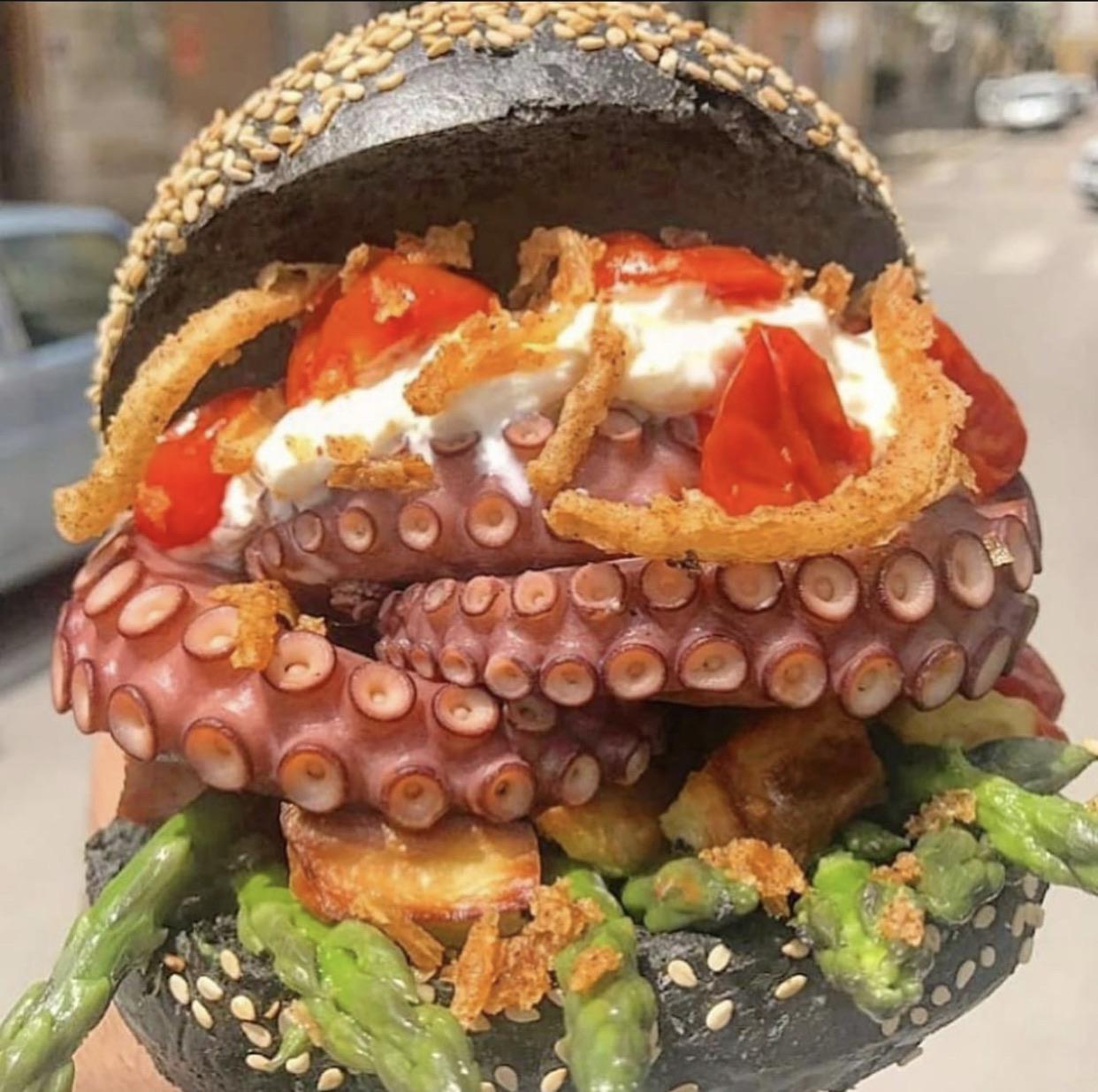 A black sesame burger bun filled with octopus tentacles, fried onions, tomatoes, asparagus, diced potatoes, and creamy sauce, held in hand on a city street.