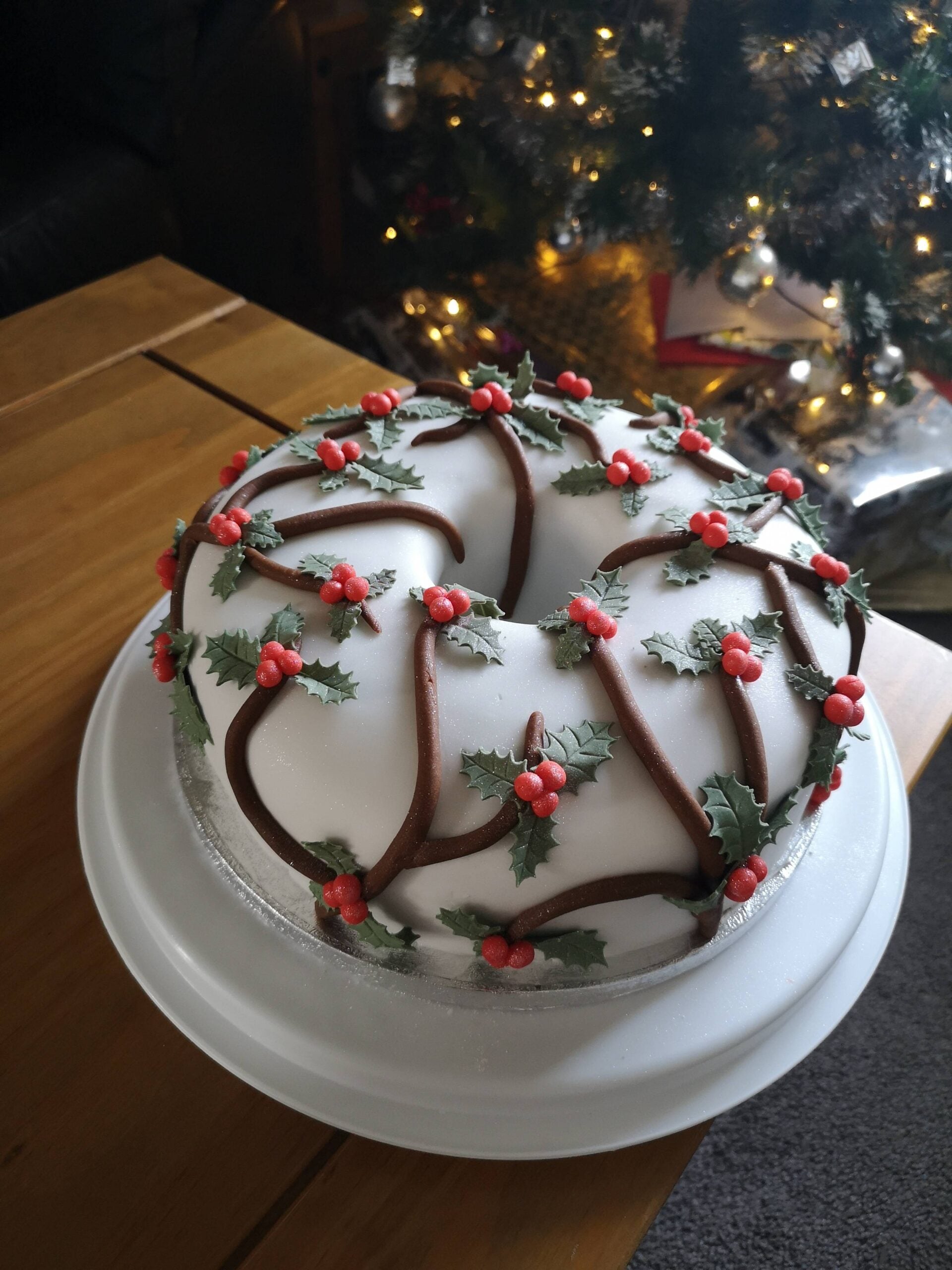 A round cake covered in white icing, decorated with holly leaves and red berries, sits on a white plate. A Christmas tree with lights is visible in the background.