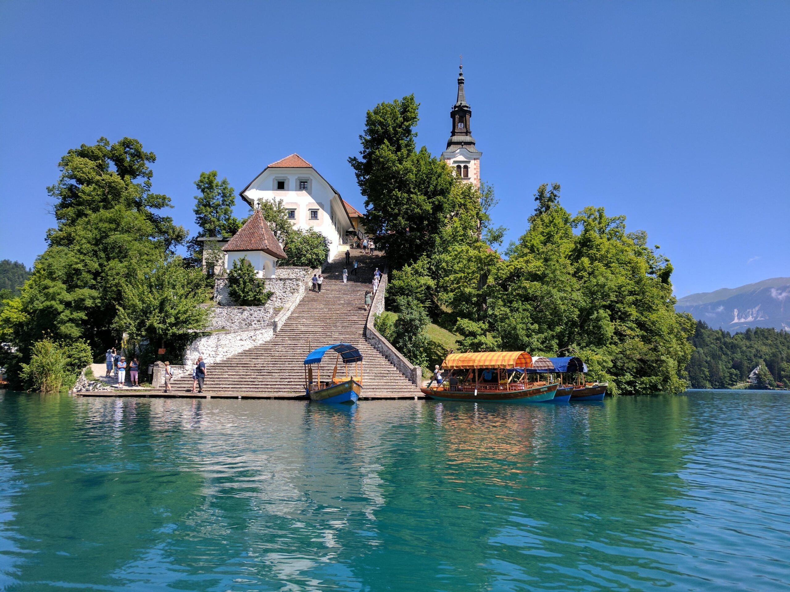 A church with a tall tower sits atop stone steps surrounded by trees on an island. Colorful boats are docked at the edge of clear turquoise water under a sunny sky.
