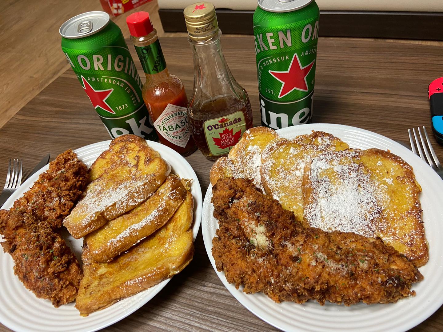 Two plates with French toast, fried chicken, and powdered sugar, next to two cans of Heineken, Tabasco, and maple syrup bottles on a wooden table.