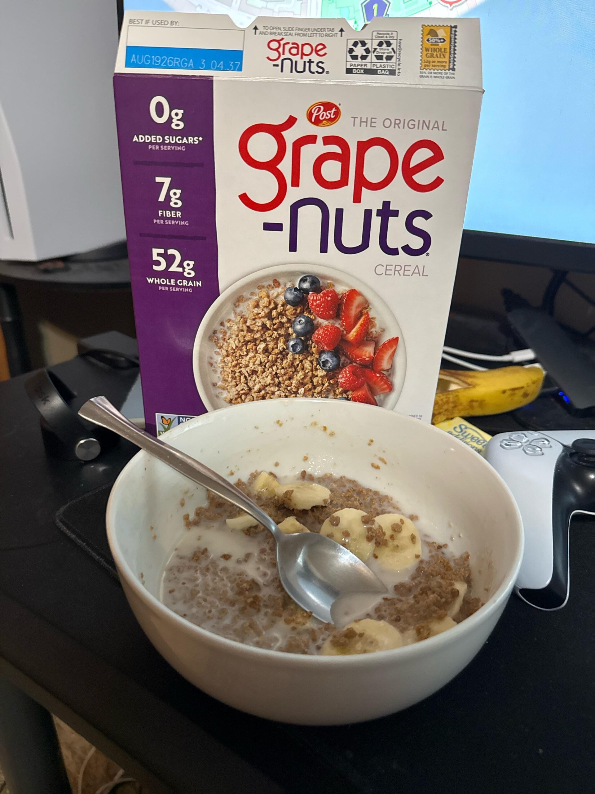 A bowl of Grape-Nuts cereal with sliced bananas and milk sits on a desk next to the cereal box. A spoon is in the bowl, and a computer mouse and keyboard are nearby.