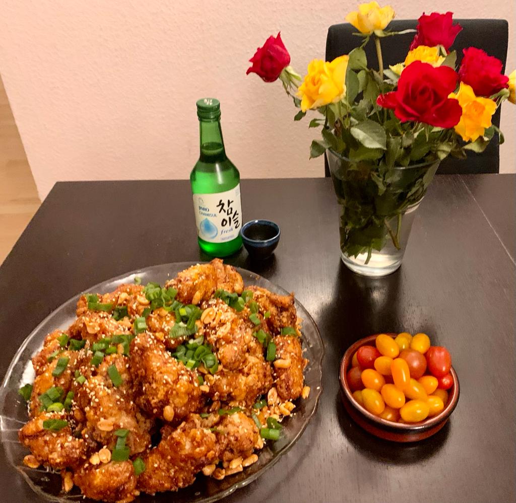 A plate of Korean fried chicken garnished with green onions and sesame seeds, a bowl of cherry tomatoes, a bottle of soju, a cup, and a vase of red and yellow roses on a dark table.