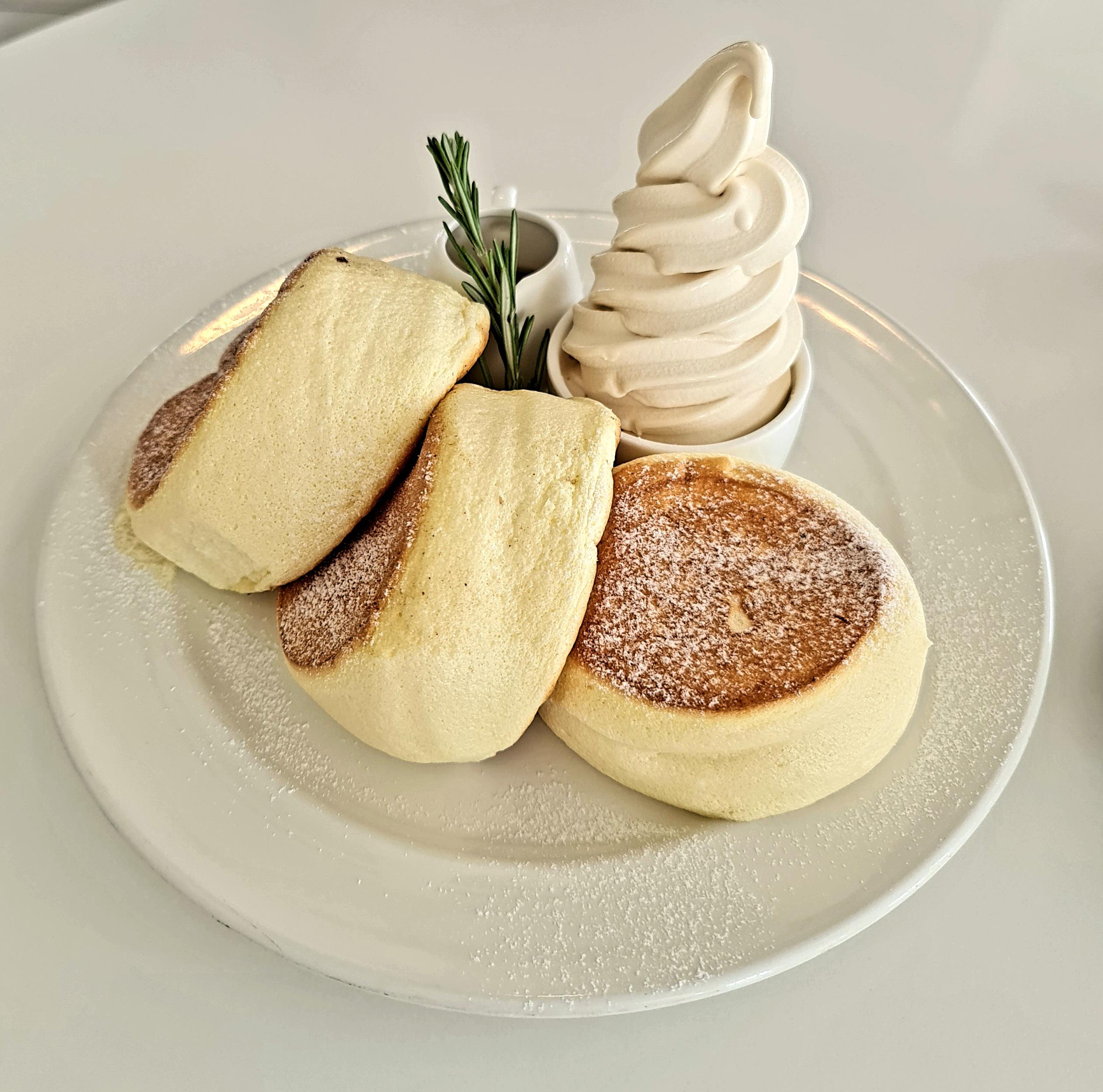 Three fluffy Japanese soufflé pancakes dusted with powdered sugar, served on a white plate with a swirl of soft-serve ice cream and a small garnish of rosemary.