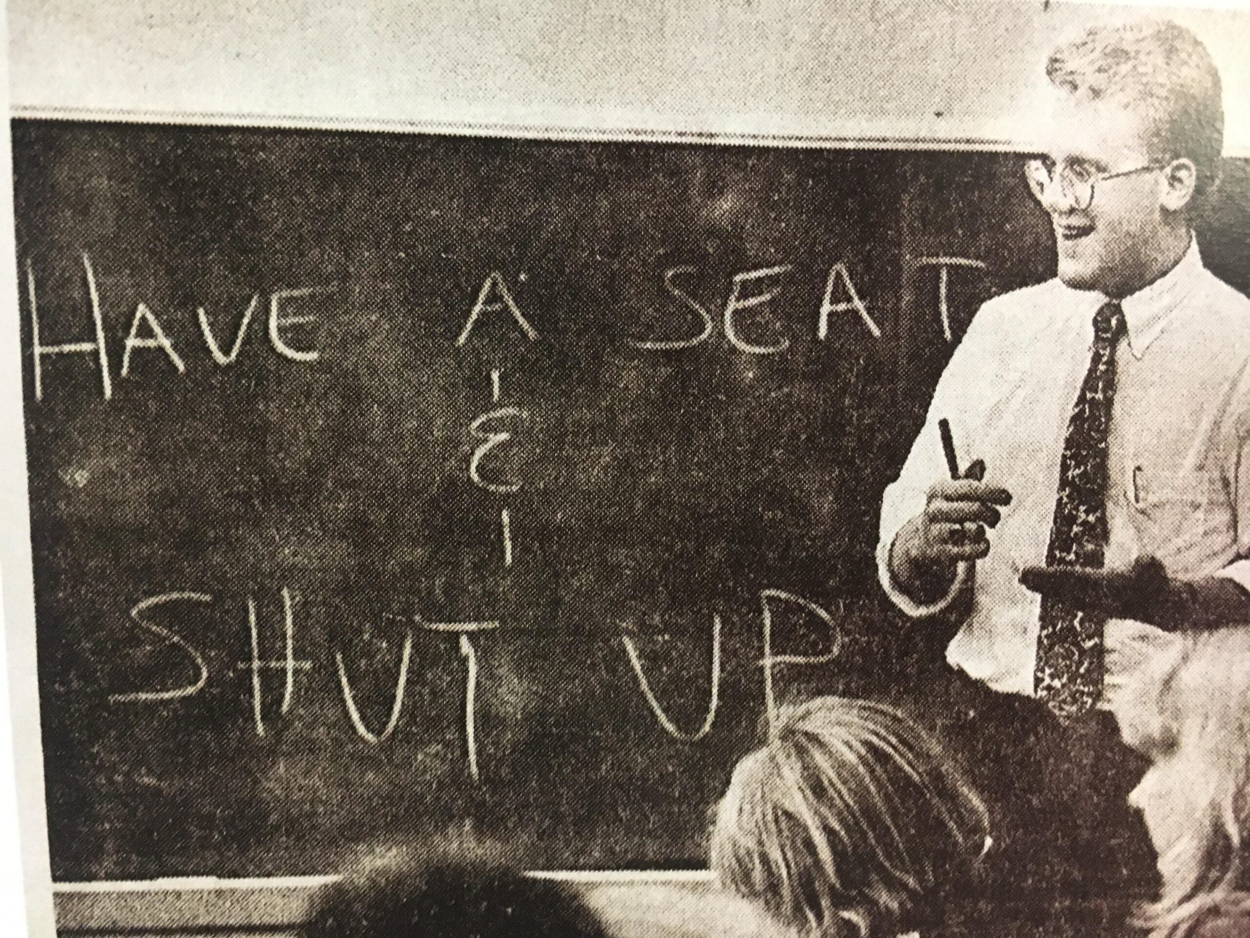 A teacher stands by a chalkboard with the words “HAVE A SEAT & SHUT UP” written on it, addressing a classroom of students.