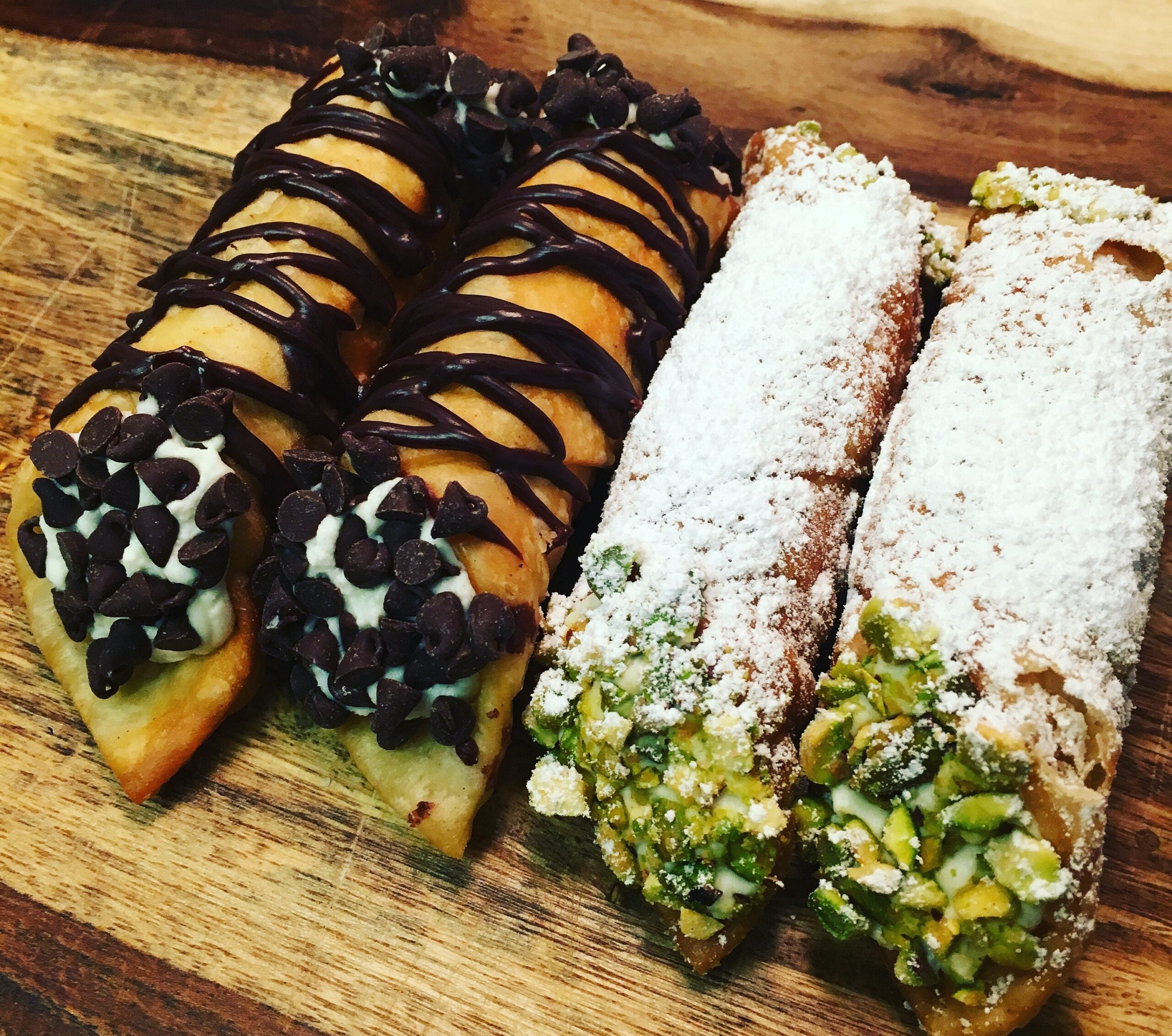Four homemade cannolis on a wooden board: two topped with chocolate chips and drizzled chocolate, and two dusted with powdered sugar and coated with chopped pistachios at the ends.
