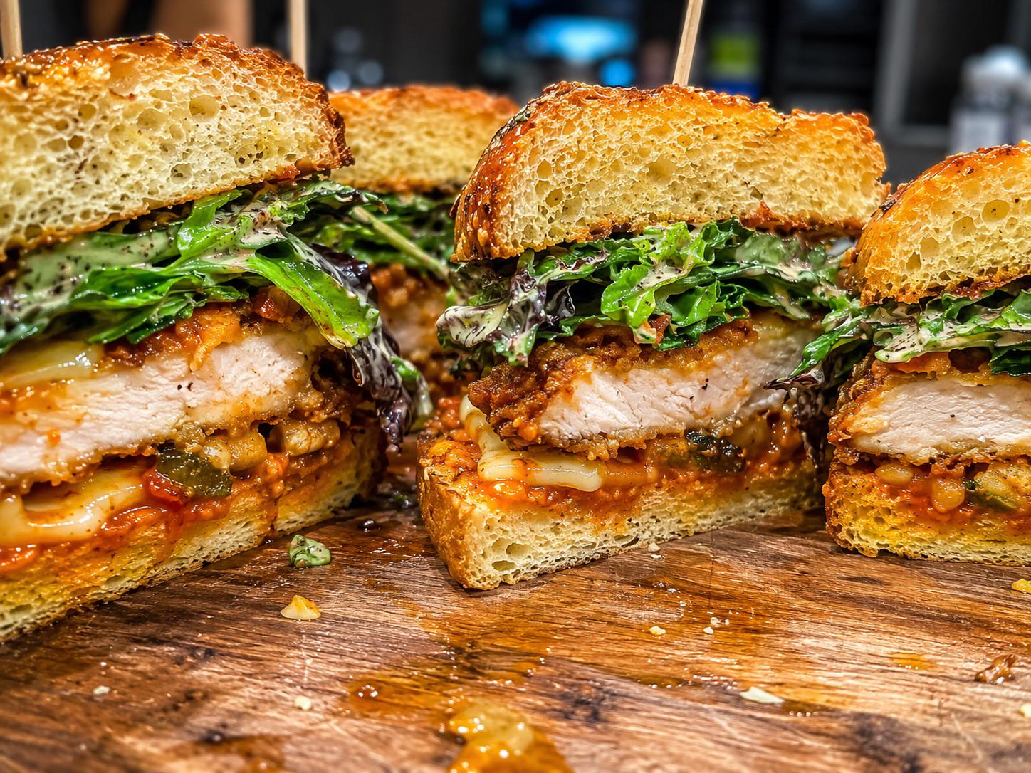 Close-up of thick sandwiches on a wooden board, stuffed with crispy fried chicken, fresh leafy greens, melted cheese, and a savory tomato-based sauce, all layered between slices of toasted bread.