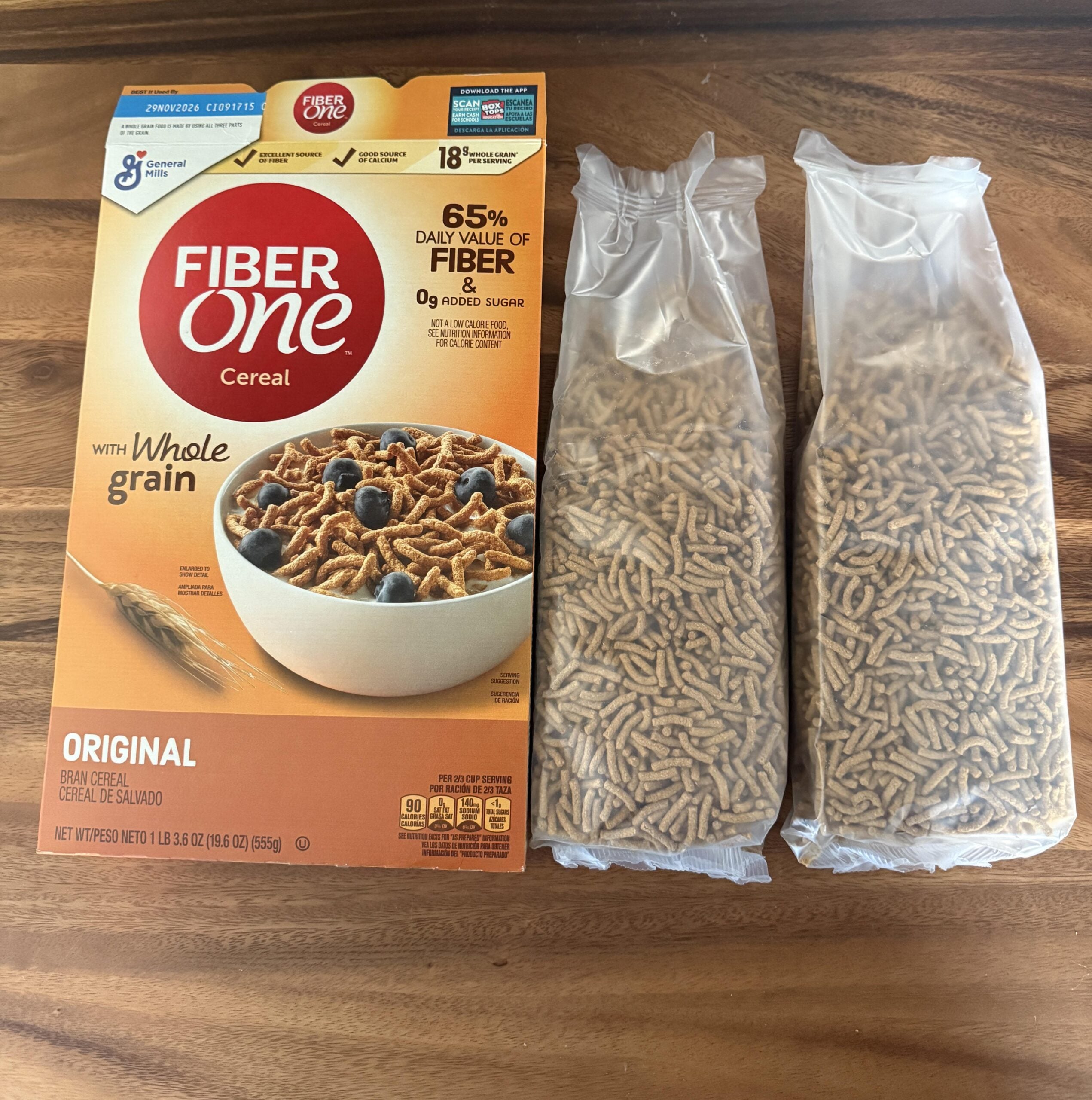 A box of Fiber One Original Cereal is on a wooden table next to two clear plastic bags filled with the cereal. The box highlights its high fiber content and whole grain ingredients.