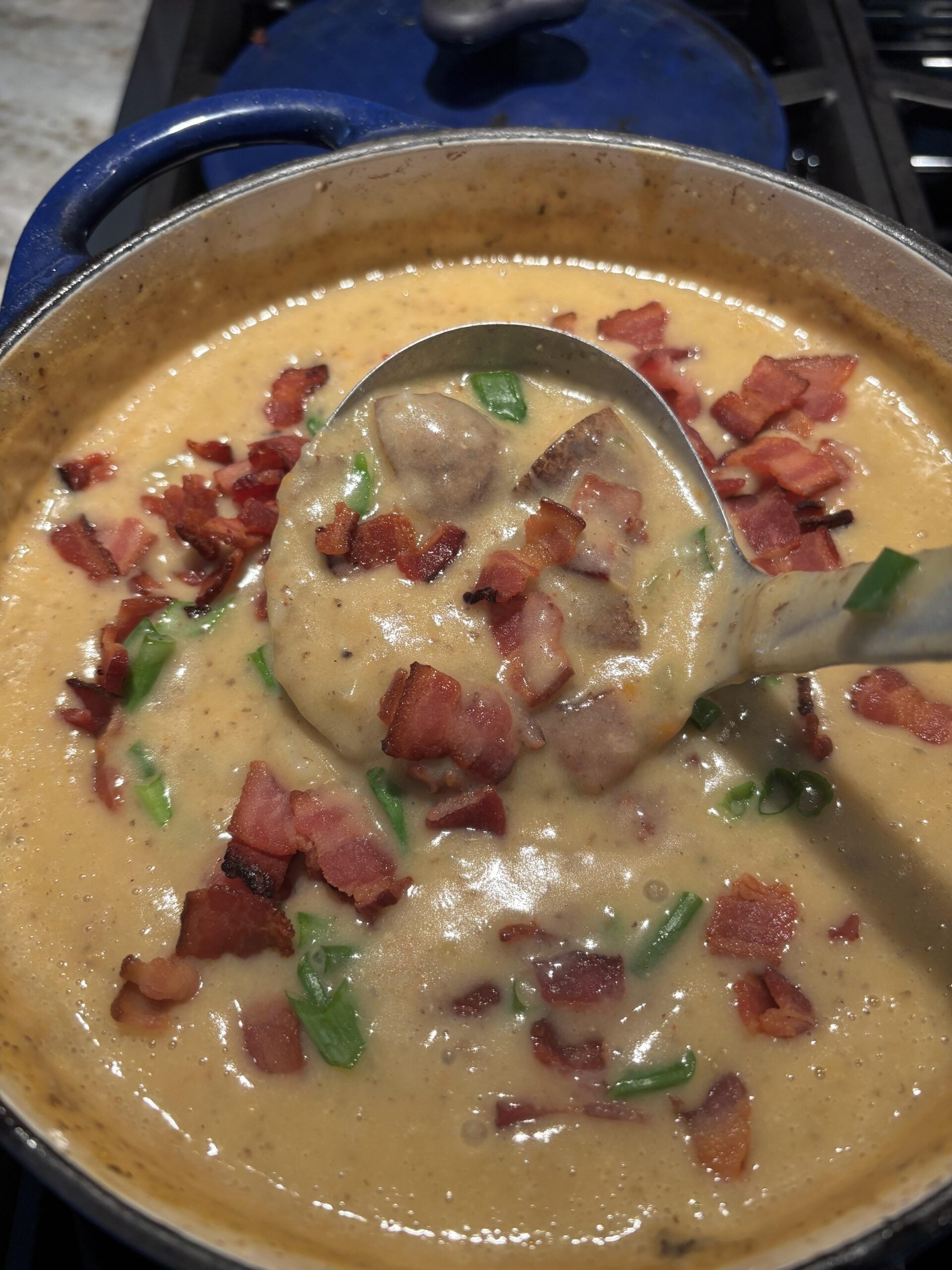 A ladle lifts creamy soup with chunks of sausage, green onions, and crispy bacon bits from a pot on the stove.