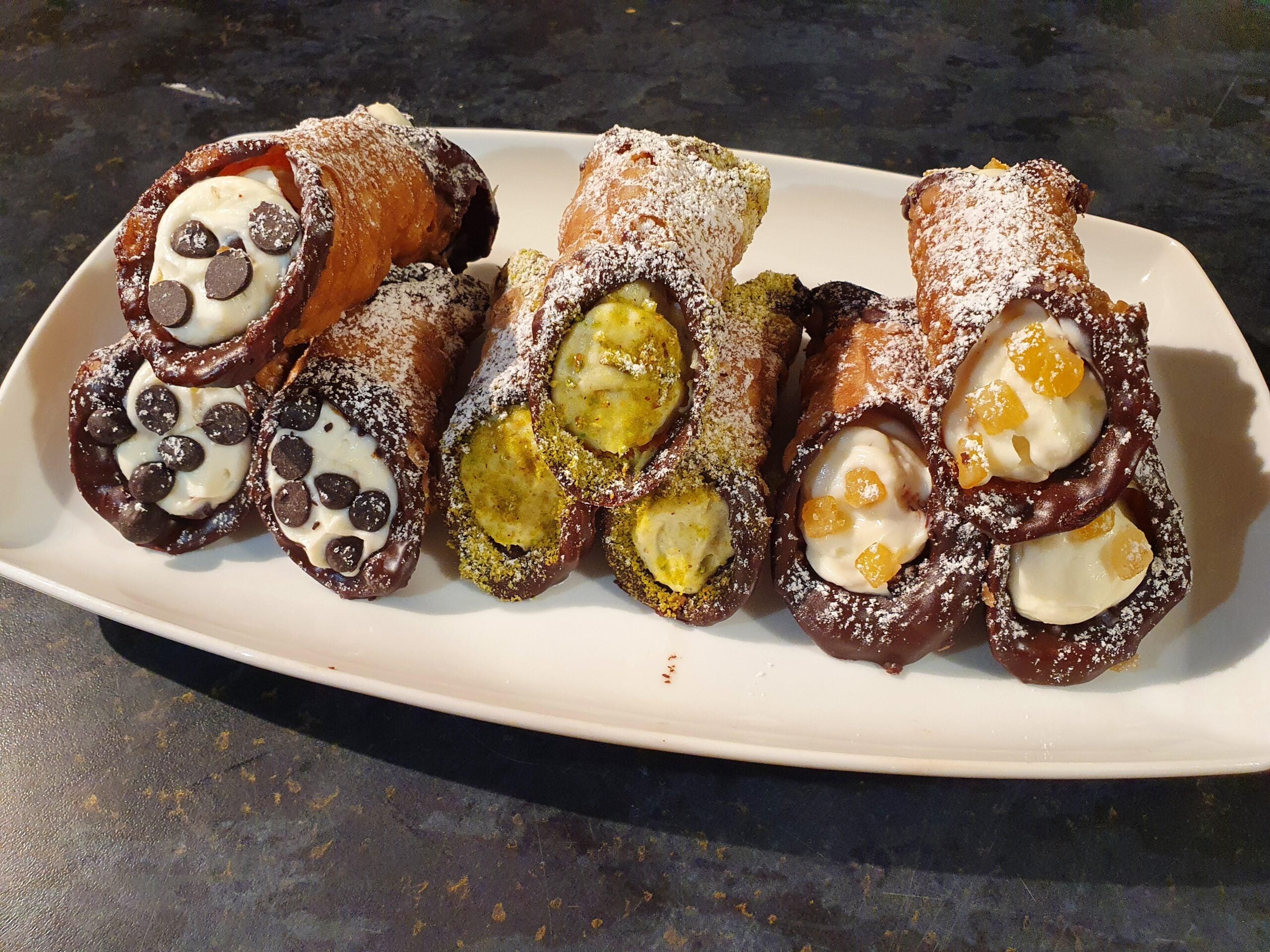 A plate of six Homemade Cannolis: two with chocolate chips, two with chopped pistachios, and two with candied orange pieces. All are dusted with powdered sugar and have chocolate-dipped ends.
