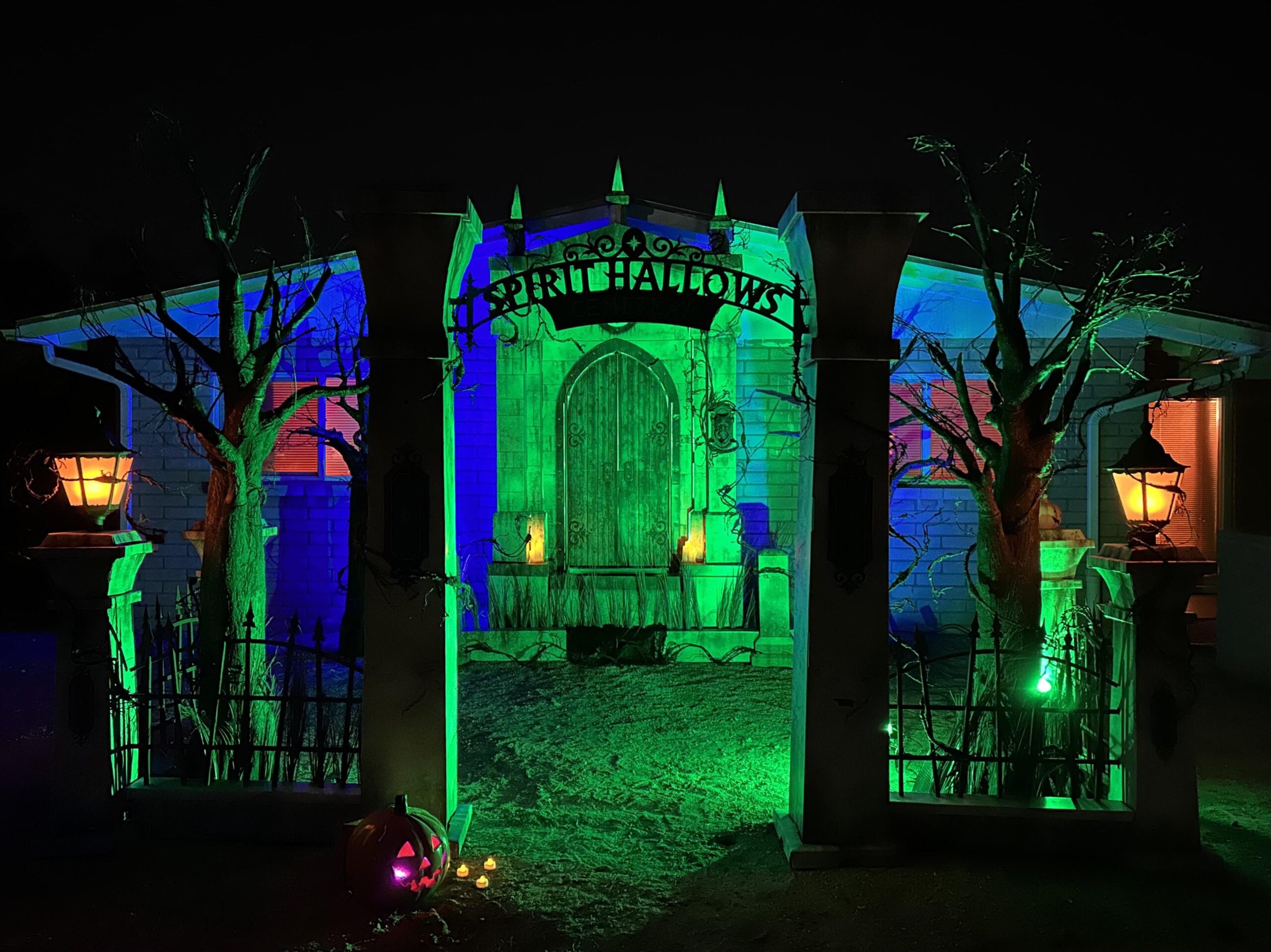 A spooky, dimly lit Halloween house with a sign reading “Spirit Hallows,” eerie green and blue lighting, leafless trees, lanterns, and a carved pumpkin at the entrance sets the perfect scene for unforgettable Halloween yard decorations.
