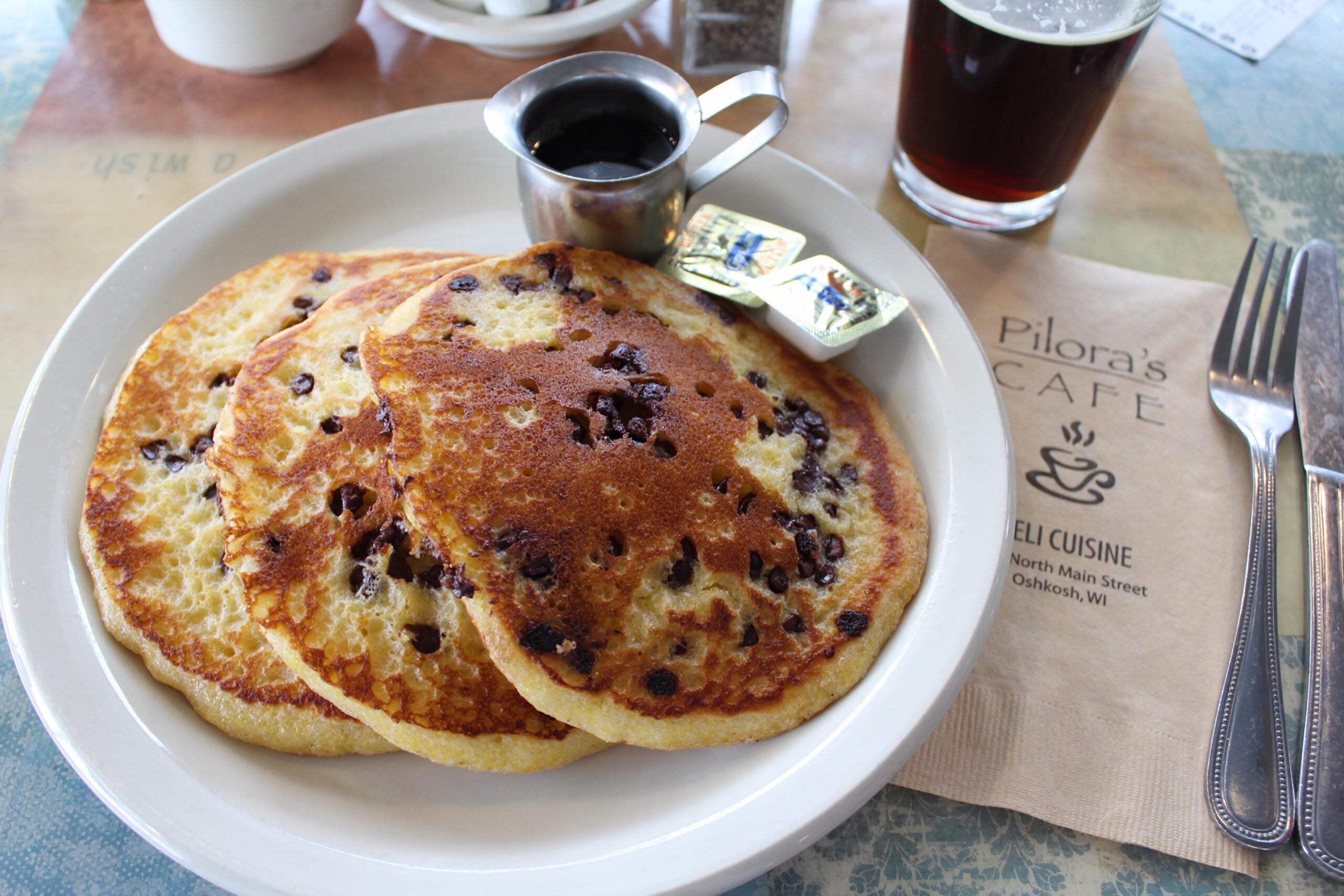 Three chocolate chip pancakes on a white plate with butter, syrup, a glass of dark beverage, and a napkin labeled “Pilora’s Cafe” on a blue table.