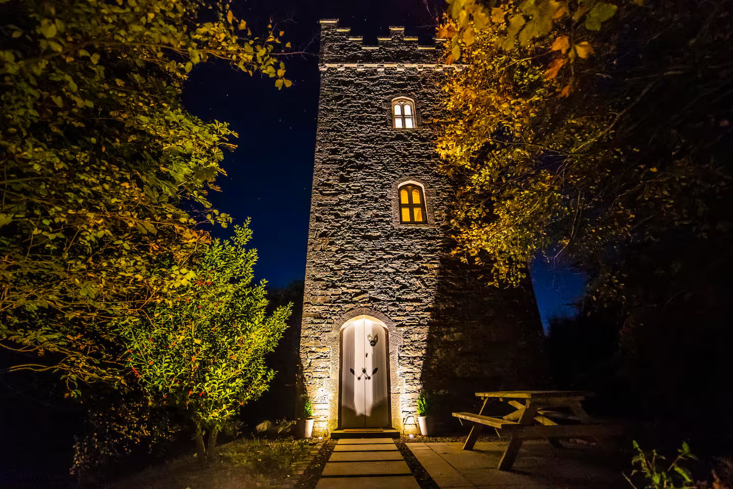 A stone tower with arched windows and a lit doorway stands at night, surrounded by trees and a picnic table under a dark, starry sky.