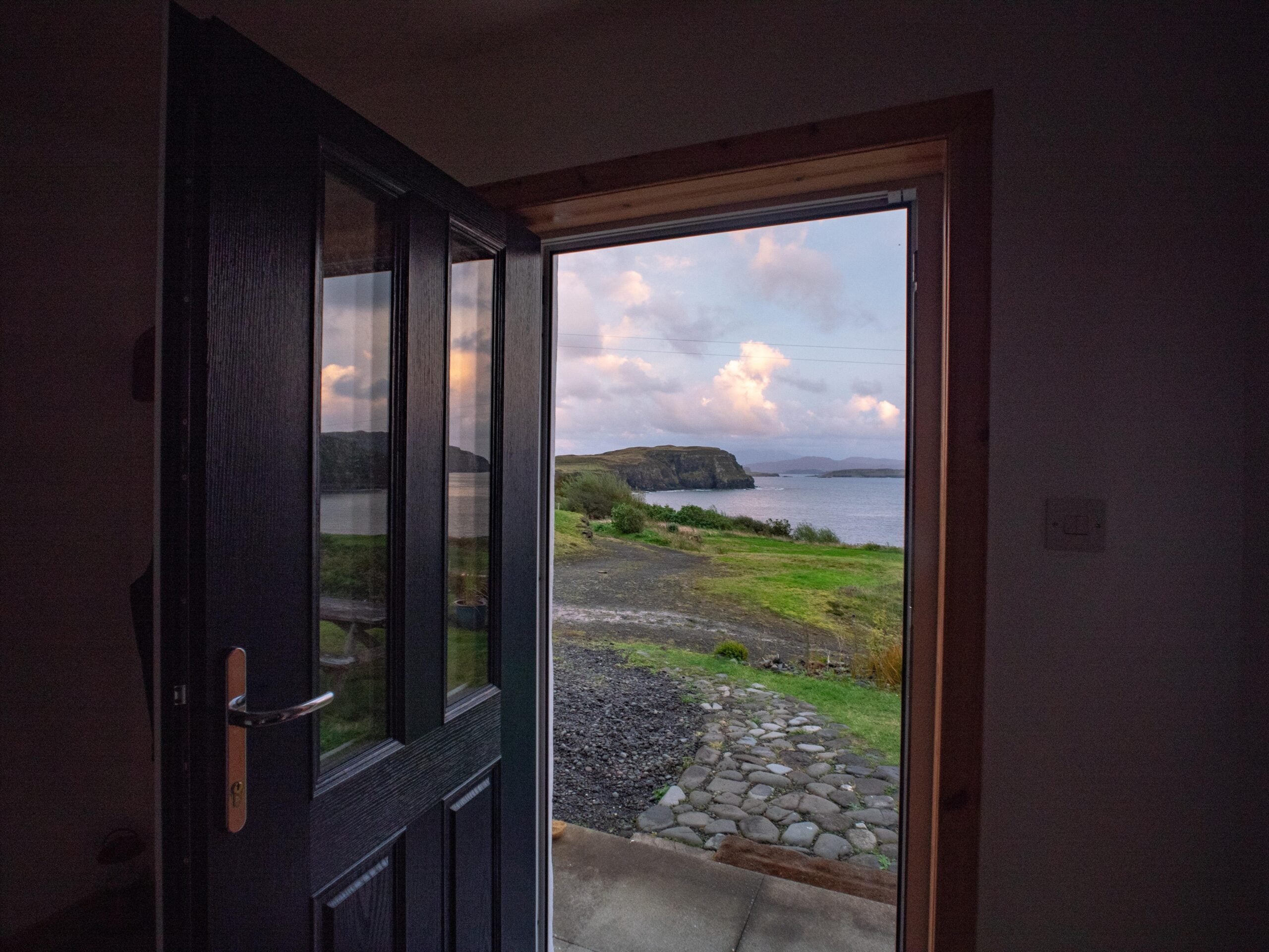 A partially open door reveals a scenic view of a grassy landscape, rocky path, and calm water with cliffs and clouds in the distance at sunset.