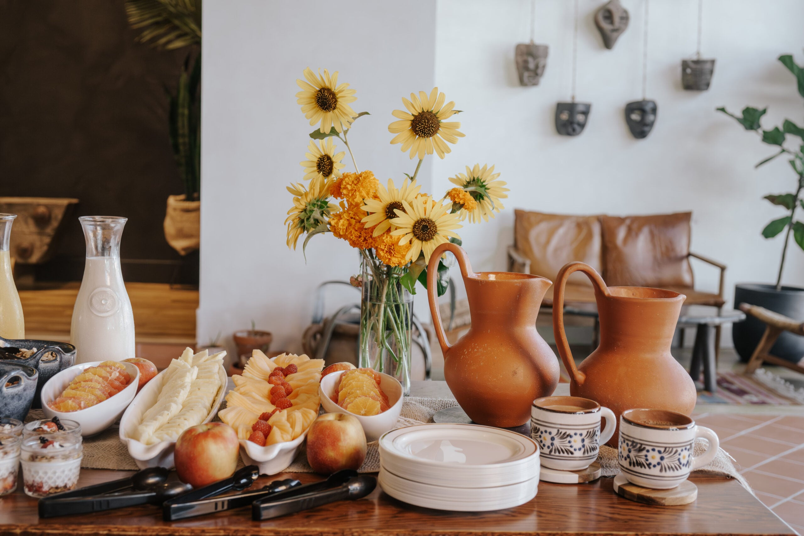 Breakfast spread with fruit, bread, apples, two jugs, cups of coffee, plates, and a vase of yellow flowers on a wooden table; cozy lounge area in the background.