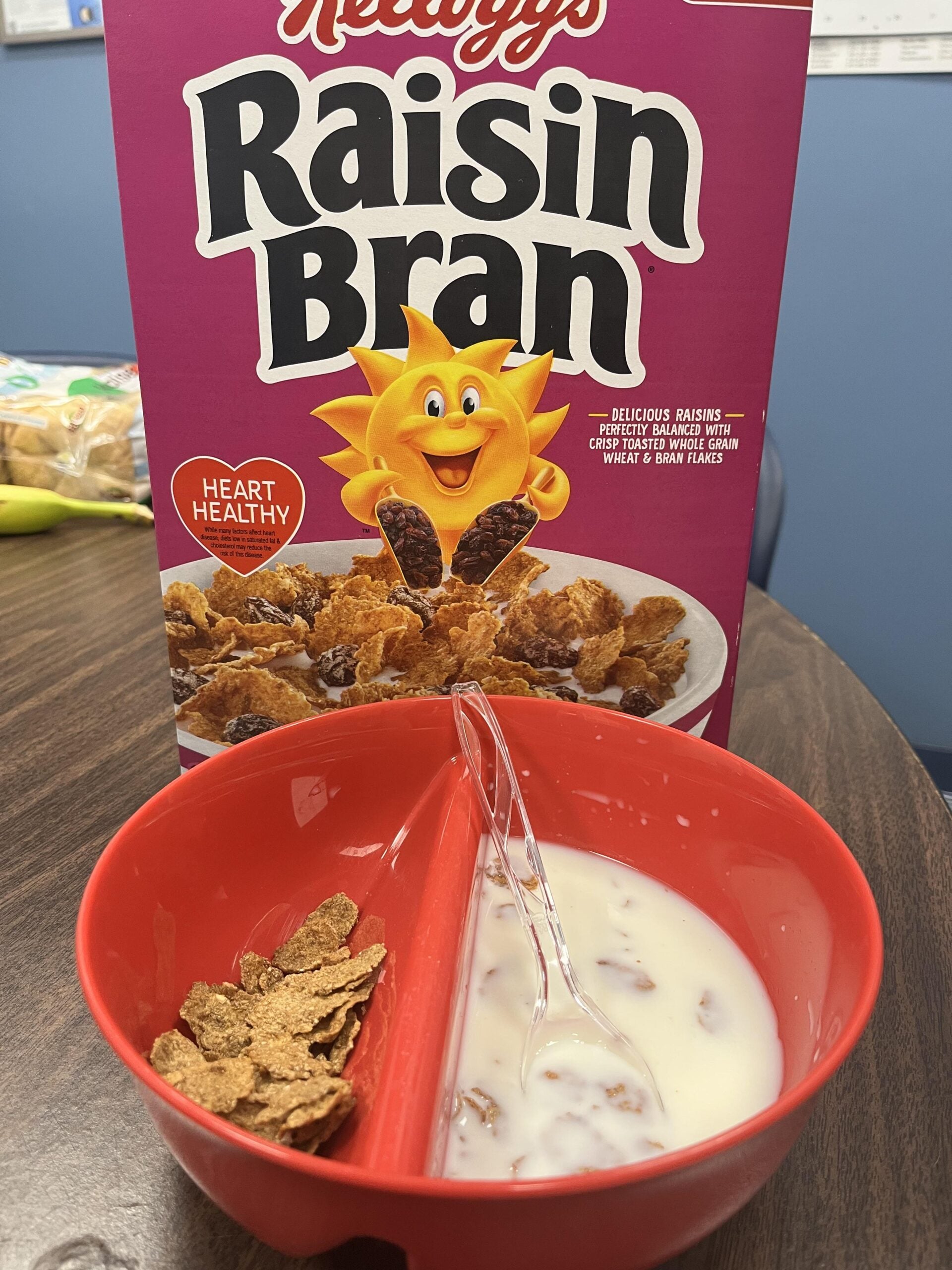 A box of Kellogg’s Raisin Bran sits behind a red divided bowl, one side holding bran flakes, the other milk. A clear spoon rests in the milk. The scene is on a wooden table.