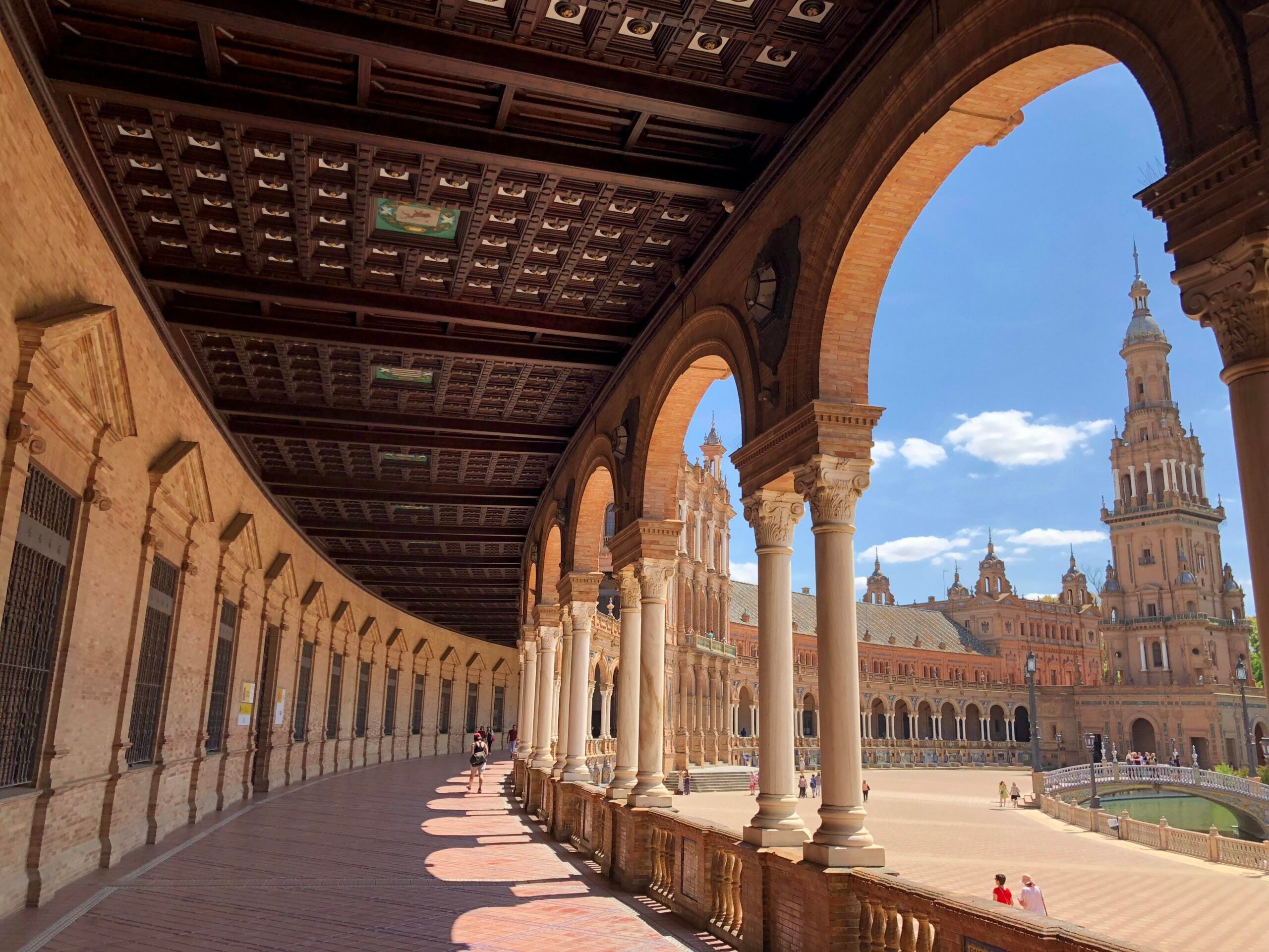 Curved arcade with arches and columns overlooks a sunlit plaza and tall historic towers under a blue sky at Plaza de España in Seville, Spain.