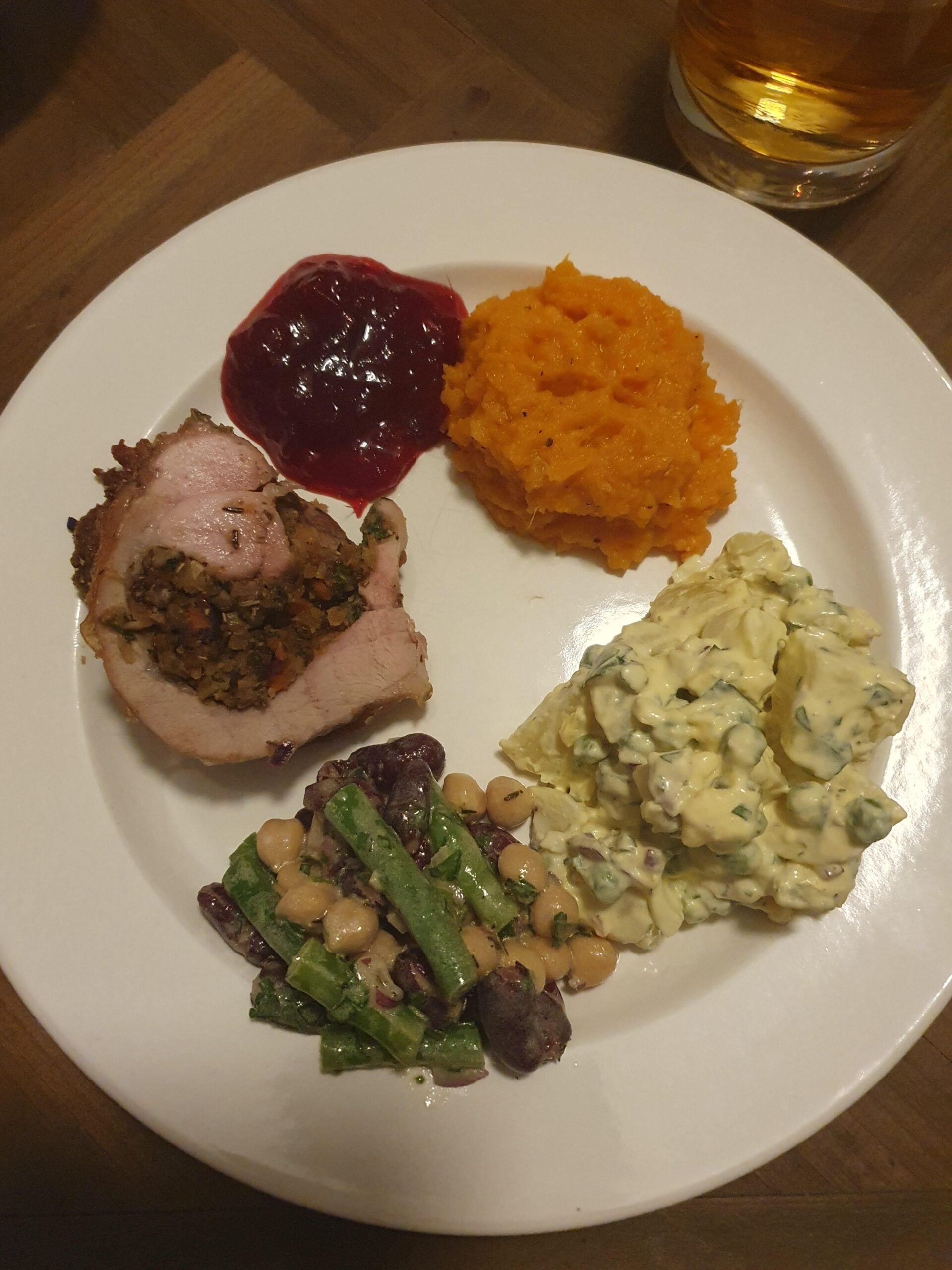 A white plate with stuffed meat, cranberry sauce, mashed sweet potatoes, potato salad, and a mixed bean salad. A glass of amber drink is visible in the background.