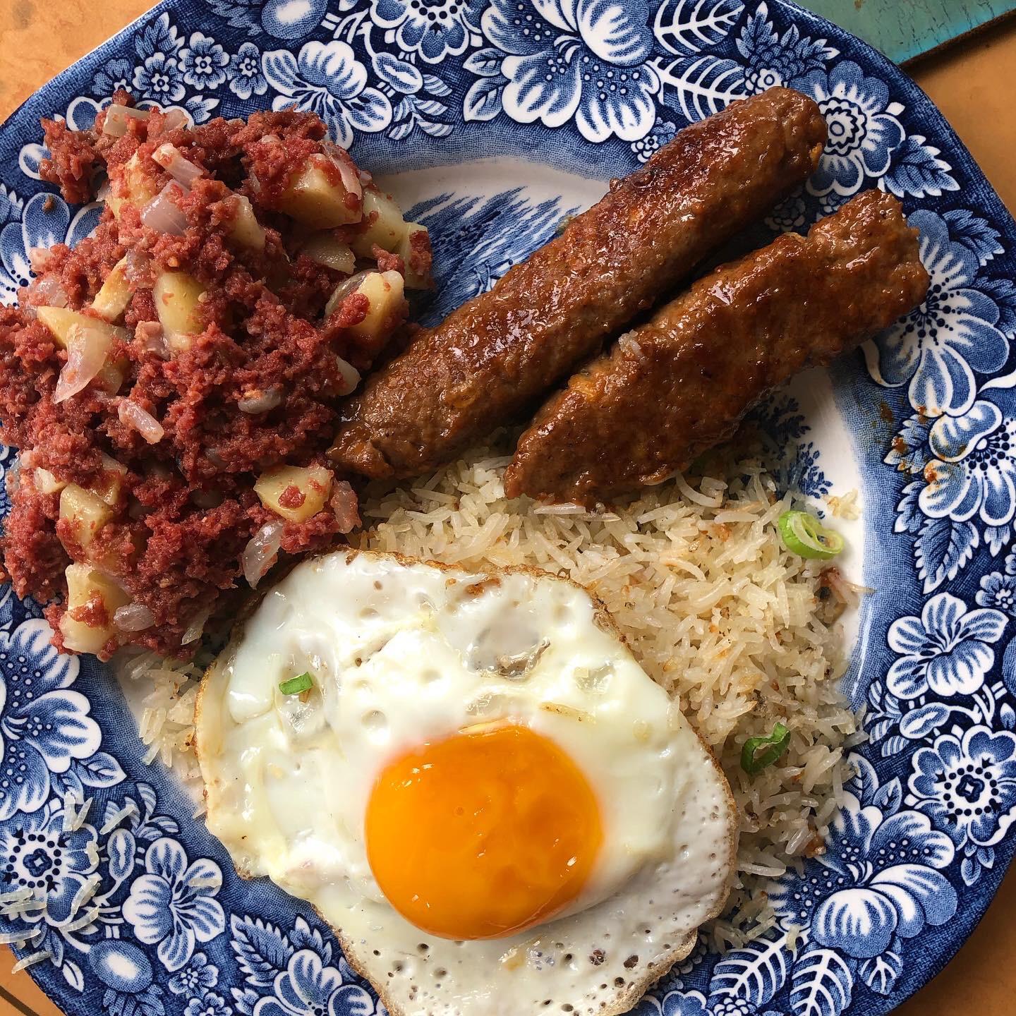 A blue and white plate with fried rice, a sunny-side-up egg, two sausage links, and corned beef hash with diced potatoes.