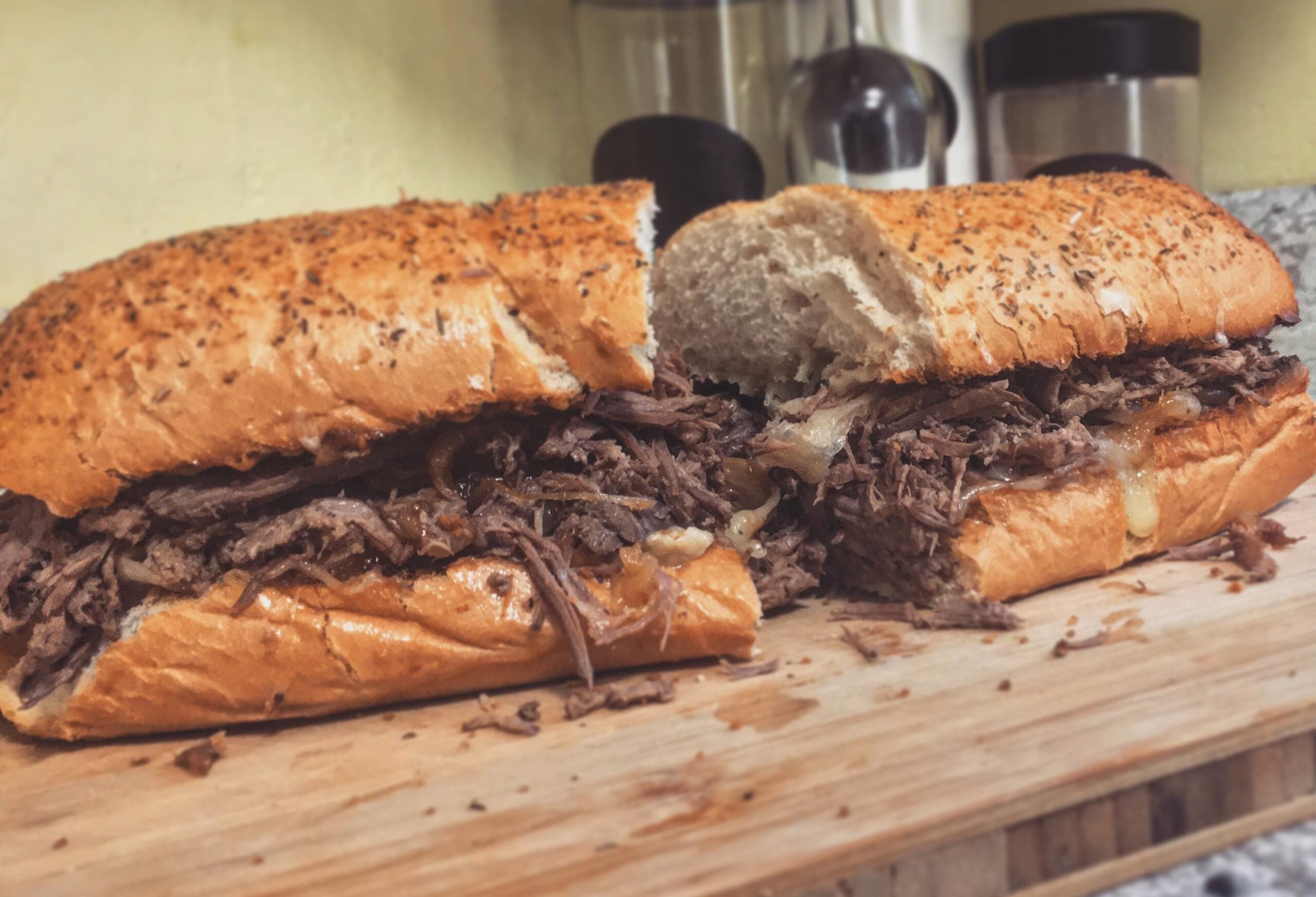 A close-up of a large sandwich on a wooden cutting board, filled with shredded beef and melted cheese, with a seasoned, toasted hoagie roll. Kitchen items are blurred in the background.