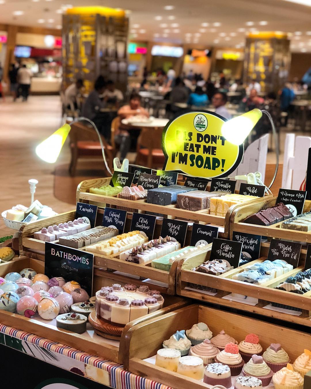 A market stall displays colorful soap bars and bath bombs arranged neatly, resembling desserts. A sign reads “Don’t eat me, I’m soap!” People are seated at tables in the blurred background.