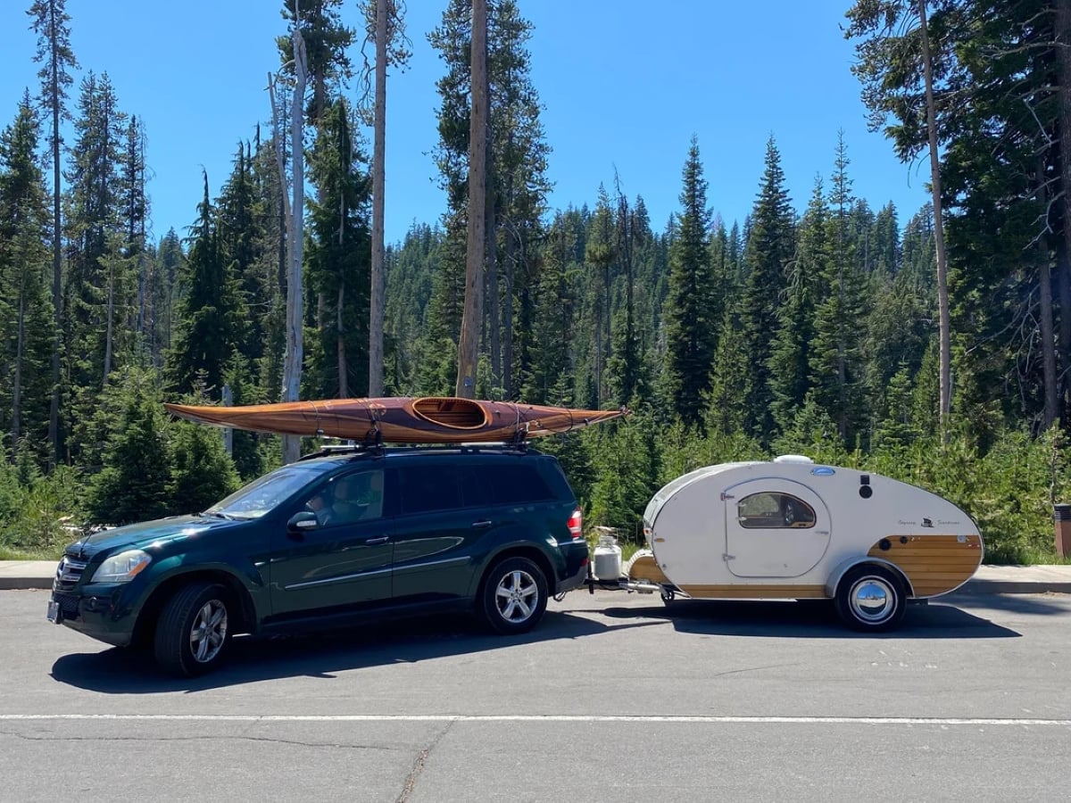 A dark SUV with a kayak on its roof is parked, towing a small teardrop camper trailer. The scene is set in a forested area with tall pine trees and clear blue sky in the background.