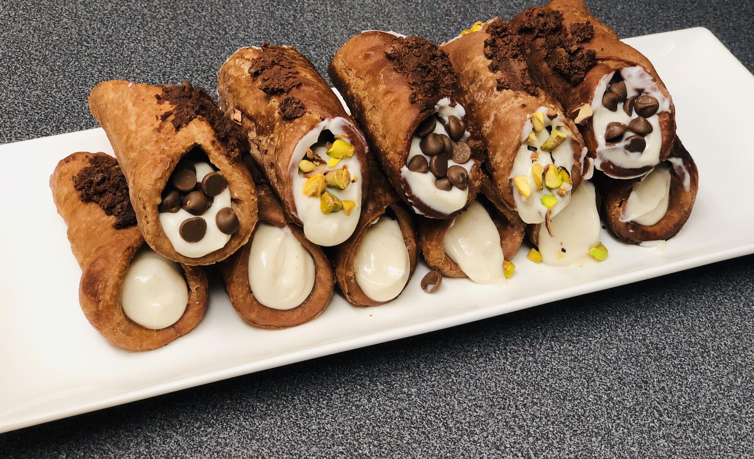A row of homemade cannolis on a white plate, filled with cream and topped with chocolate chips, crushed pistachios, and chocolate crumbles, set on a dark countertop.