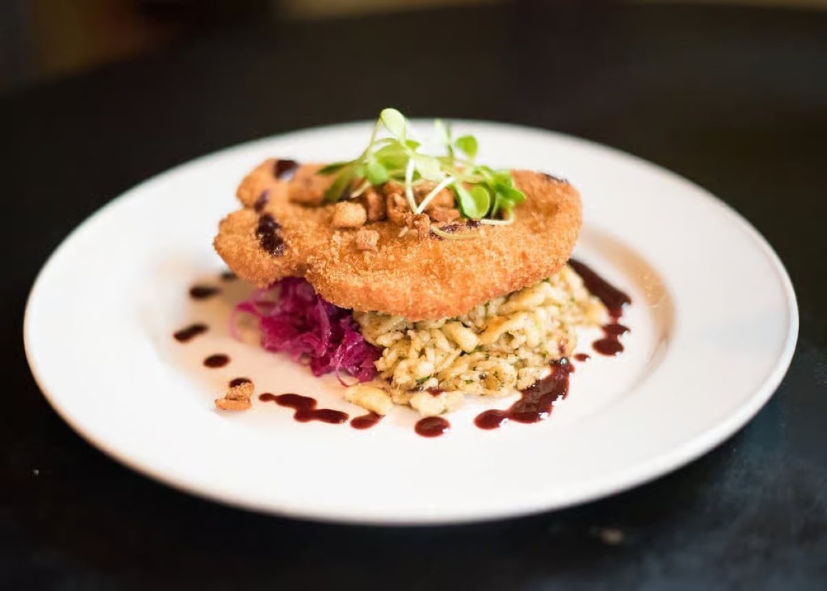 A breaded cutlet sits atop creamy noodles and red cabbage, garnished with microgreens and a dark sauce on a white plate, all on a dark table.