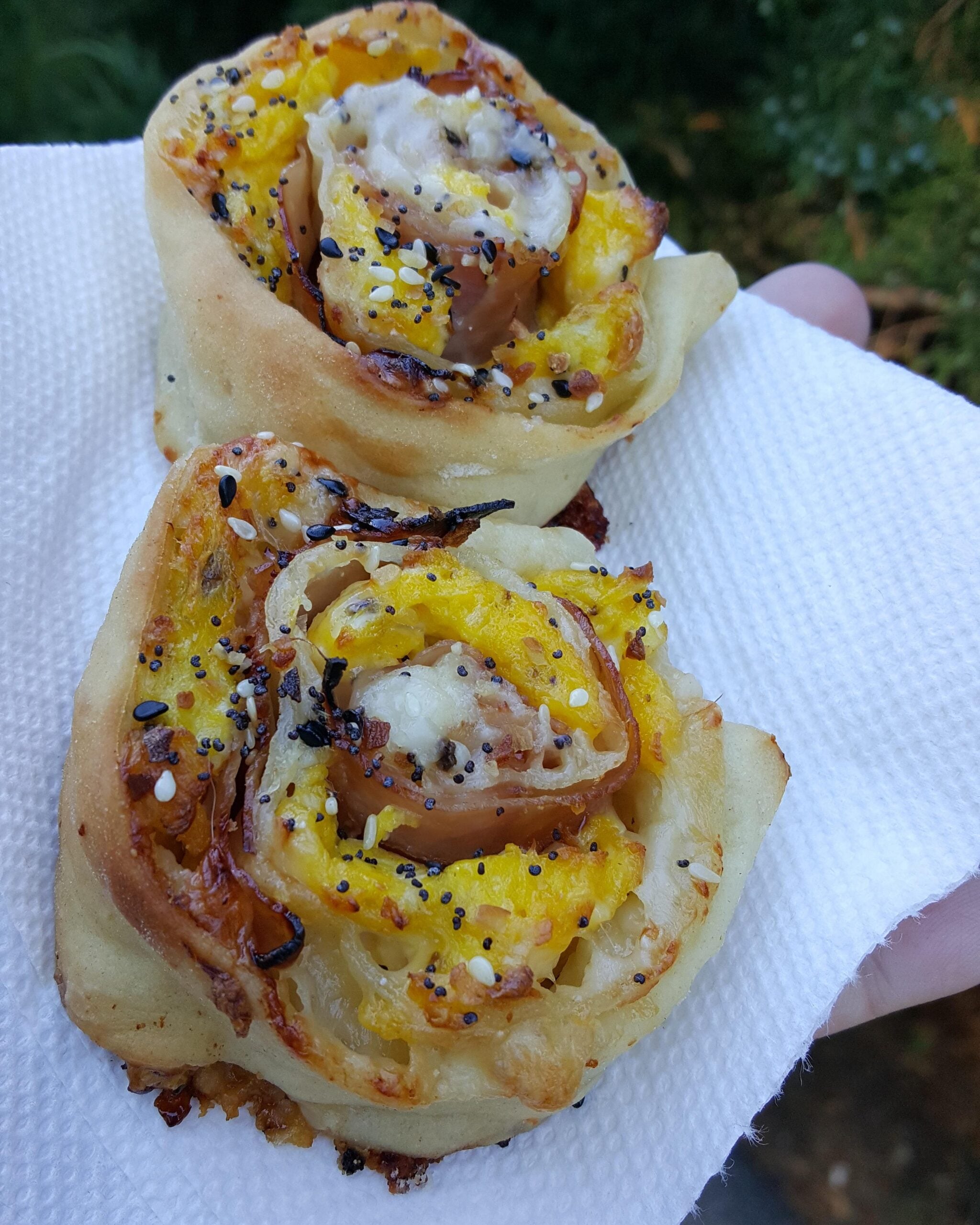 Two savory pastry pinwheels with visible poppy seeds, sesame seeds, egg, and cheese are placed on a white paper towel being held outdoors with green foliage in the background.