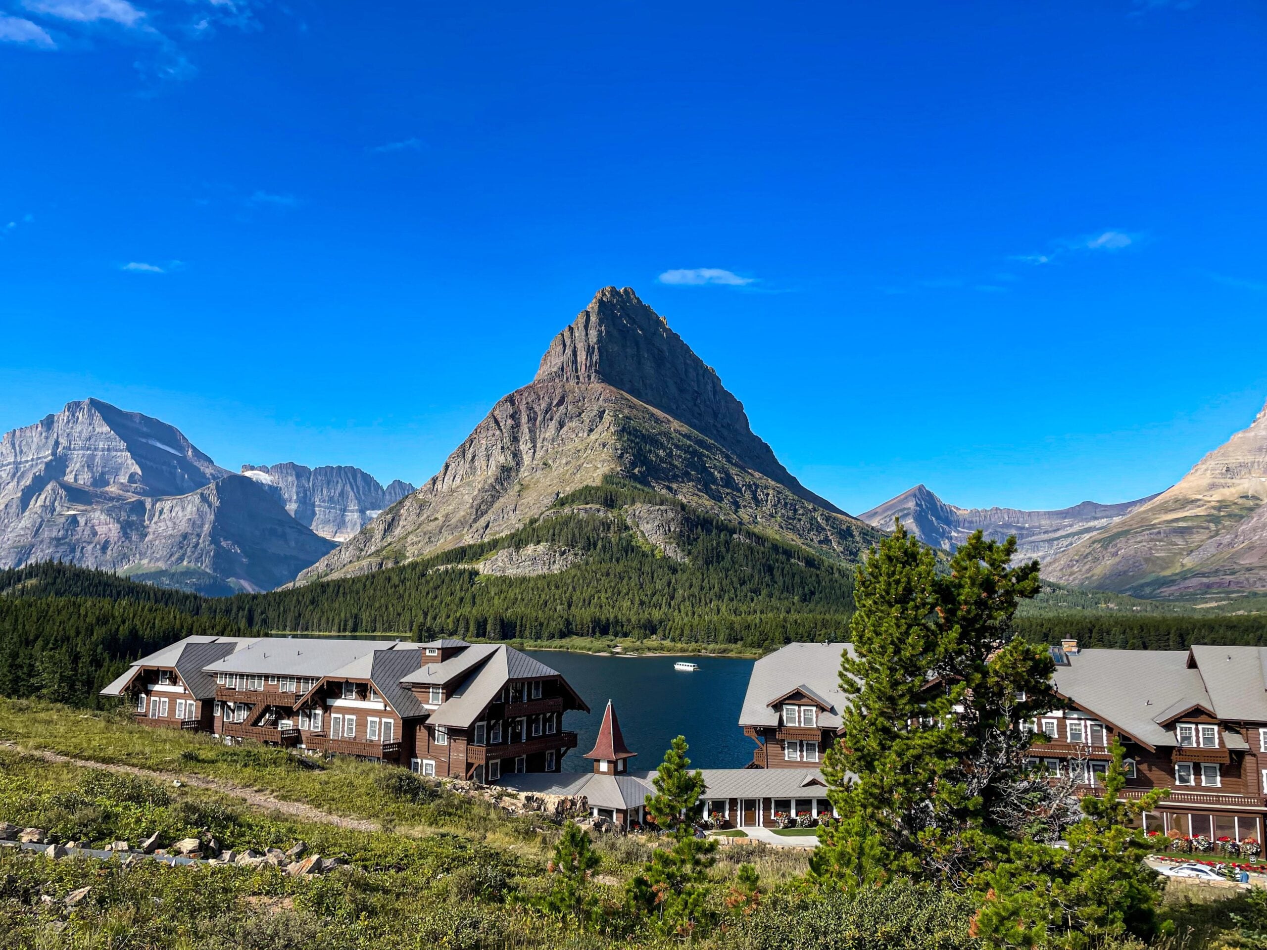 Lodge buildings beside a lake, with a tall, pointed mountain and other peaks in the background under a clear blue sky, reminiscent of the breathtaking scenery found in American National Parks.