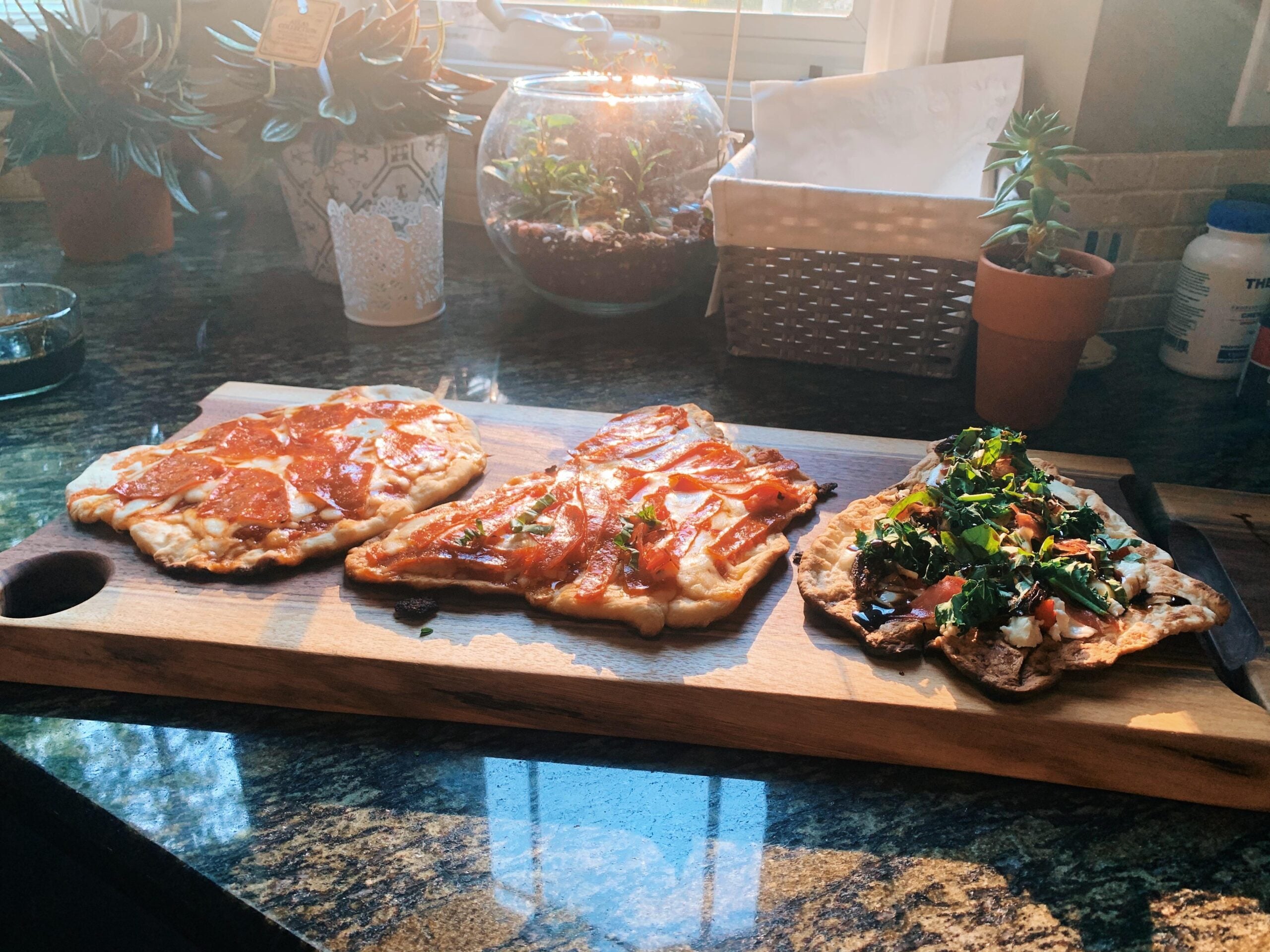 Three differently topped flatbread pizzas on a wooden board sit on a kitchen counter, with plants, a basket, and sunlight streaming in from a window in the background.