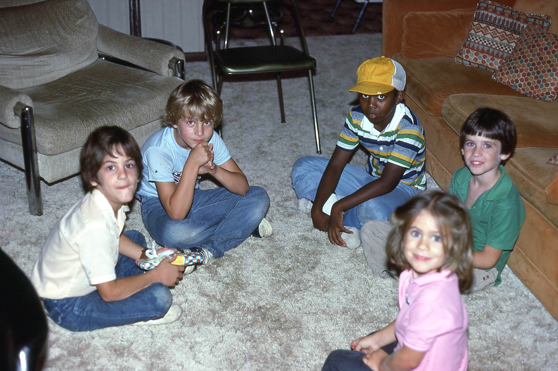 Five children sit on a beige carpet in a living room, arranged in a loose circle. They are casually dressed, some smiling, with a couch and chairs visible in the background.