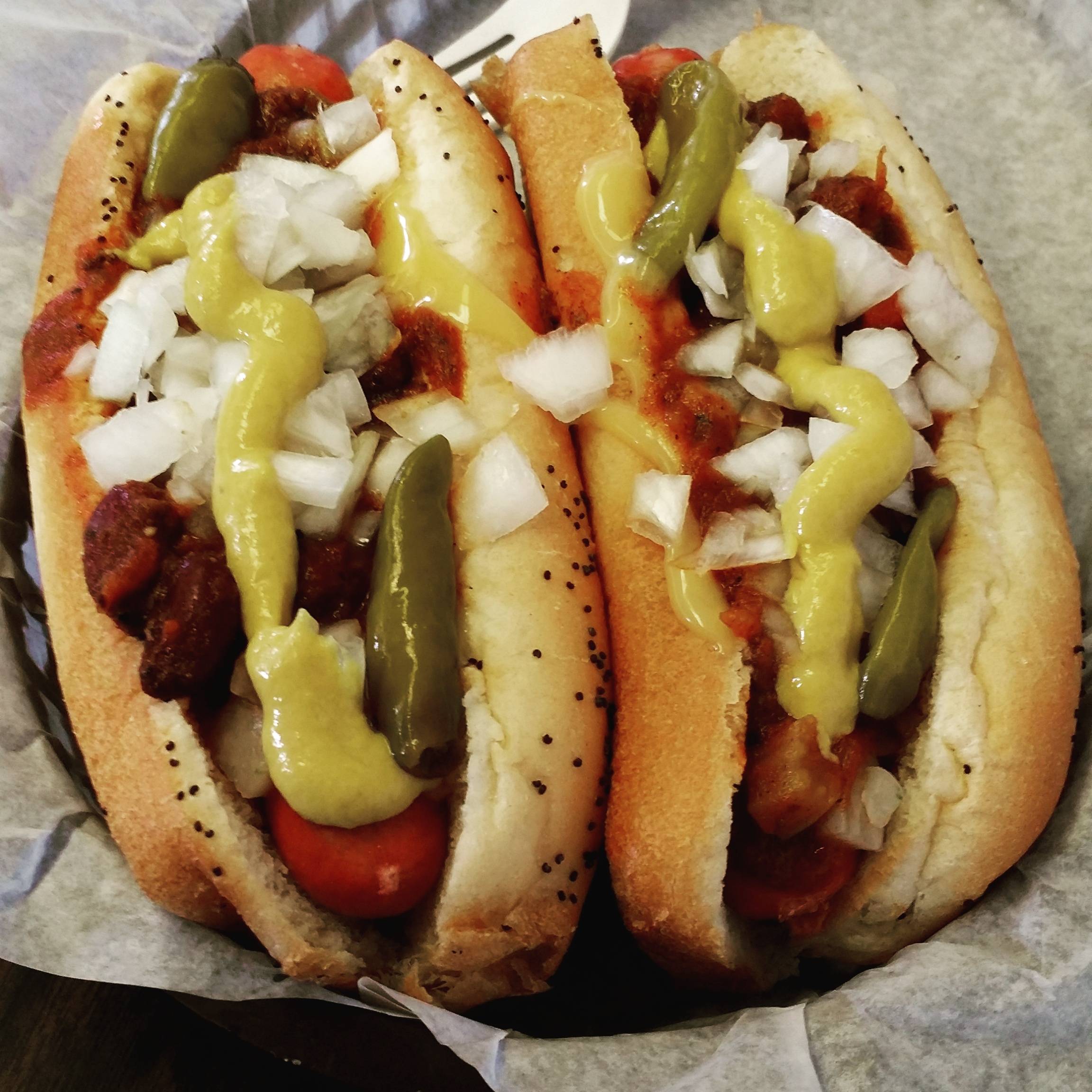 Two poppy seed buns filled with hot dogs, topped with chopped onions, yellow mustard, green peppers, and a chunky chili or meat sauce, served on parchment paper.