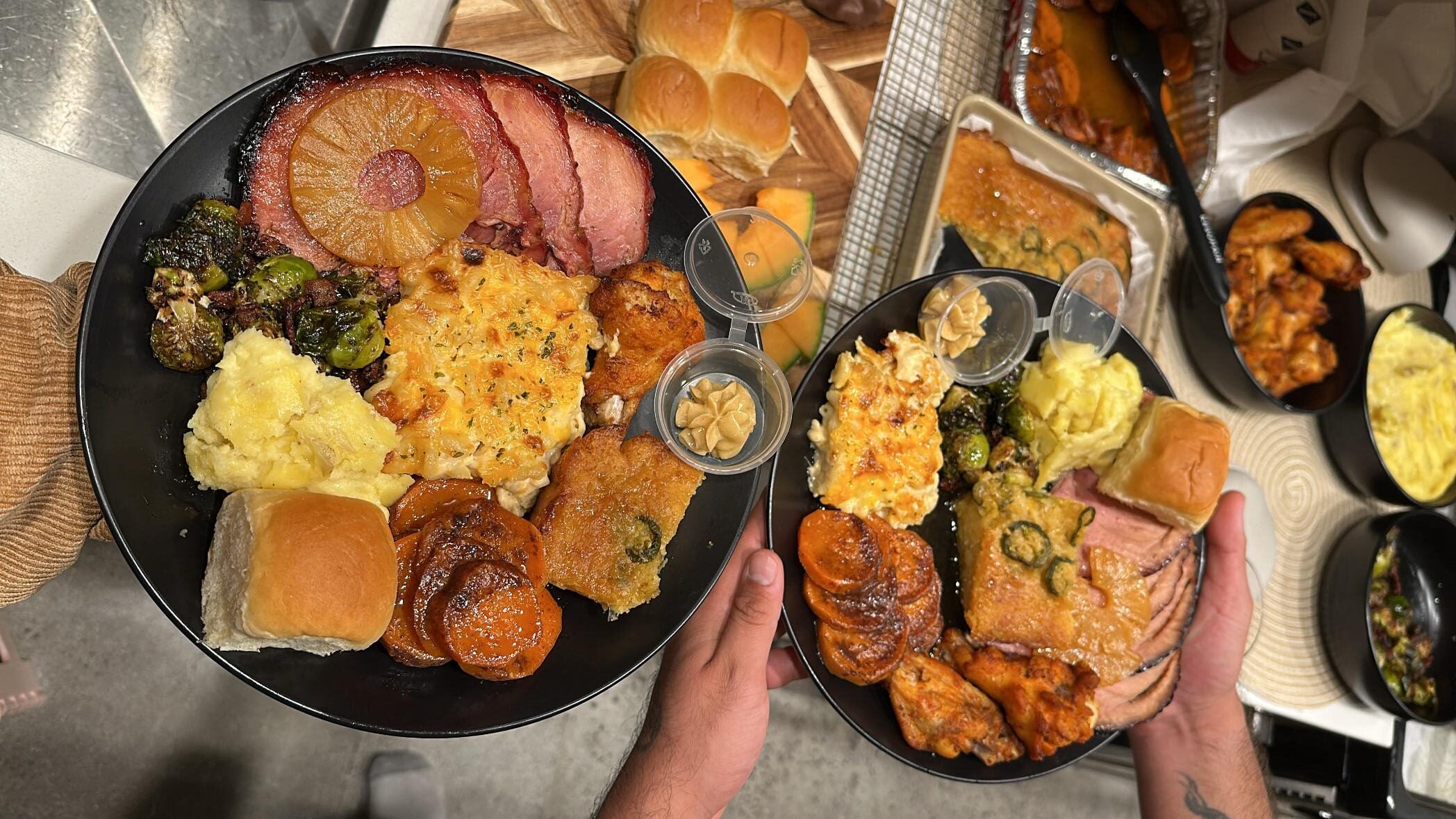 Two hands hold plates filled with ham, pineapple, mashed potatoes, rolls, sweet potatoes, casserole, chicken wings, and vegetables above a table with more food.