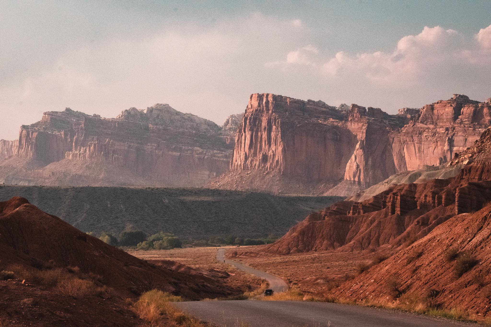 A winding road leads through a dry, rocky desert landscape with towering red rock cliffs and mesas under a partly cloudy sky.