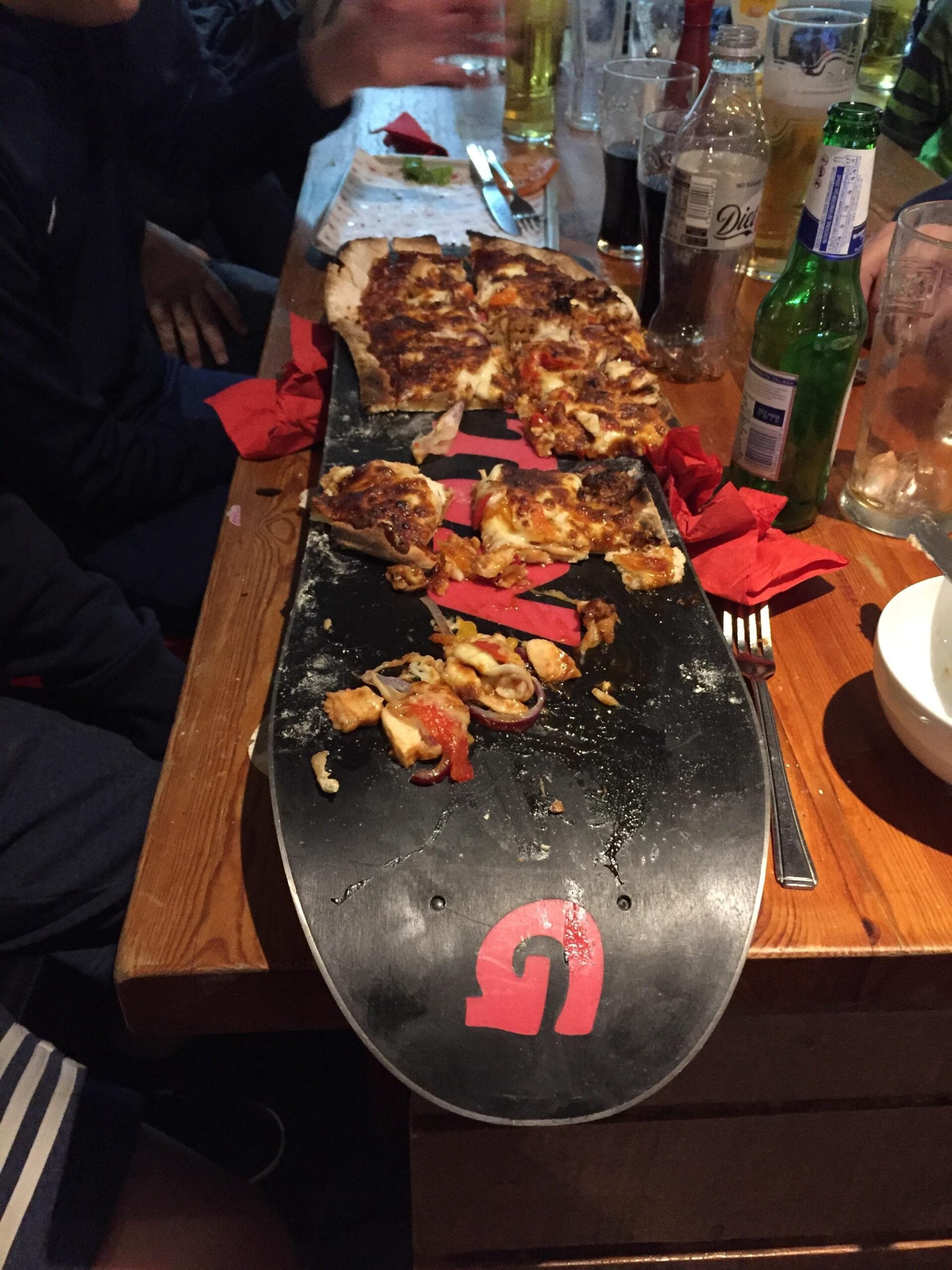 Slices of pizza are served on a skateboard deck placed on a wooden table surrounded by people, drinks, and red napkins.